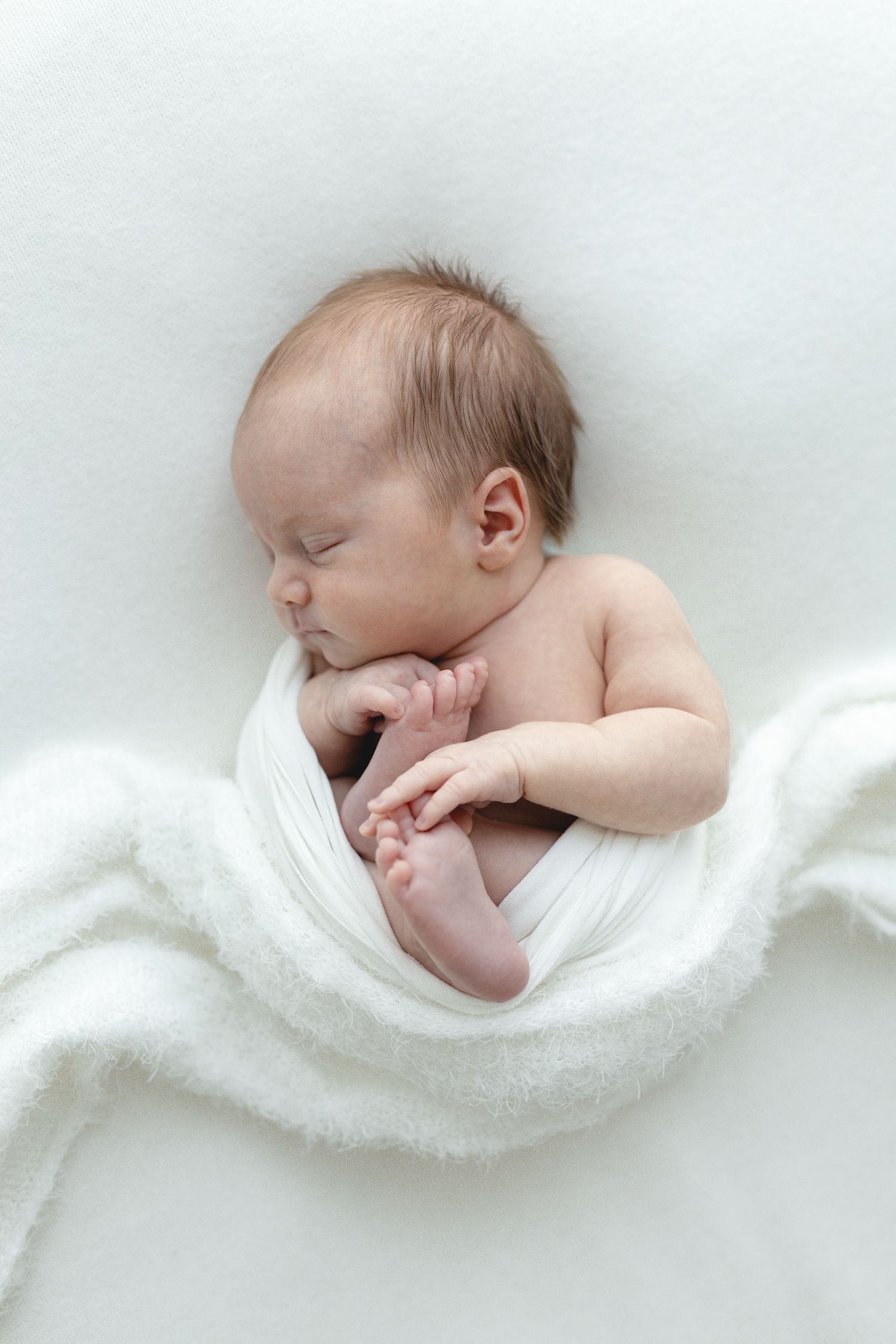 Peaceful sleeping newborn baby wrapped in soft white blanket on light fabric. Newborn sessions in studio or at home.