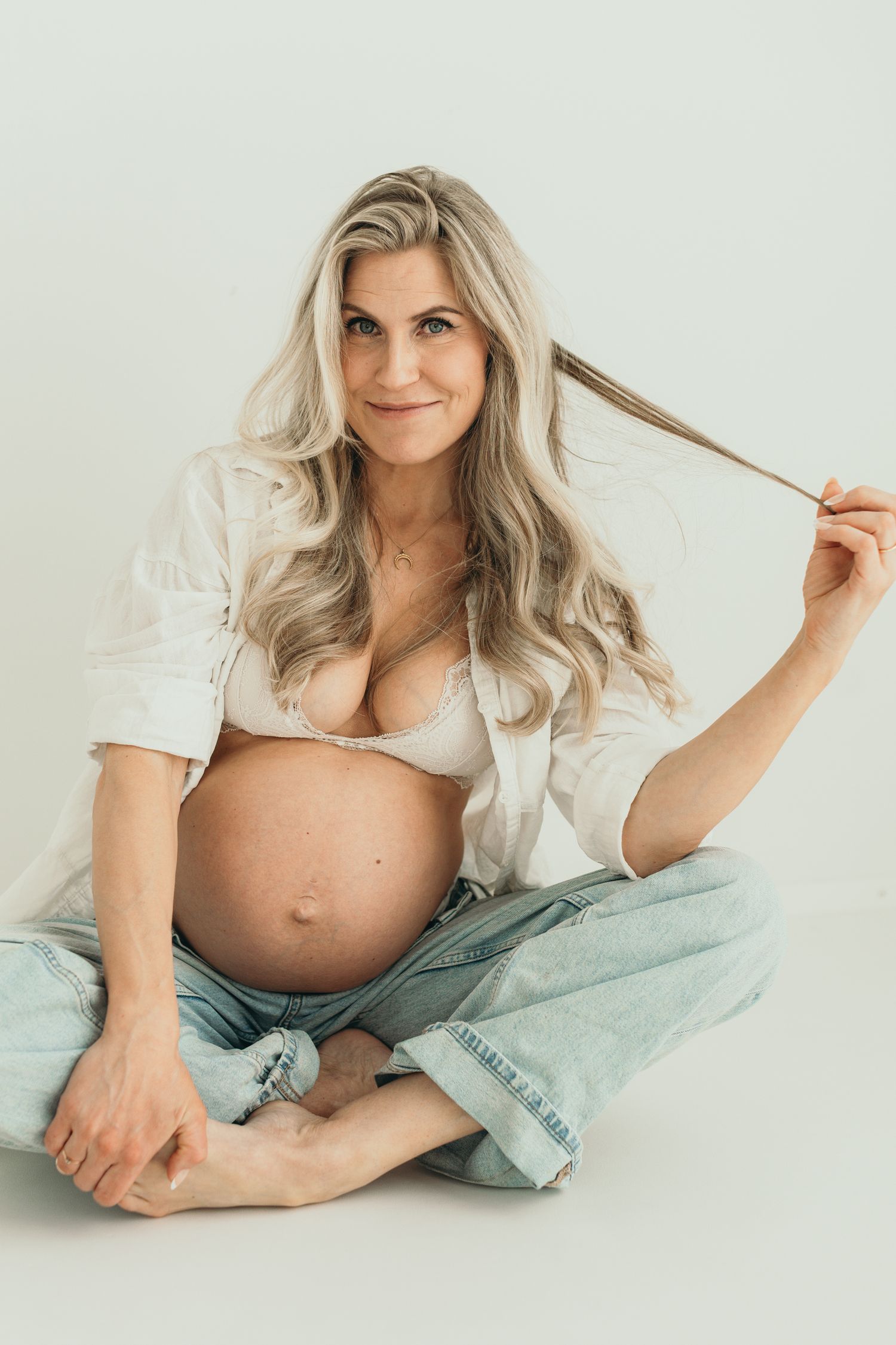 Smiling pregnant woman with long blonde hair sitting cross-legged in open white shirt and jeans. Maternity photography at the studio.