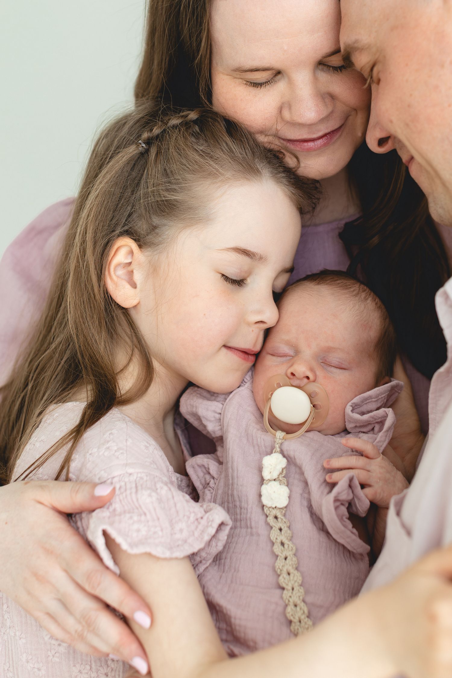 Parents and older sister tenderly embracing sleeping newborn baby with pacifier in soft pink body.