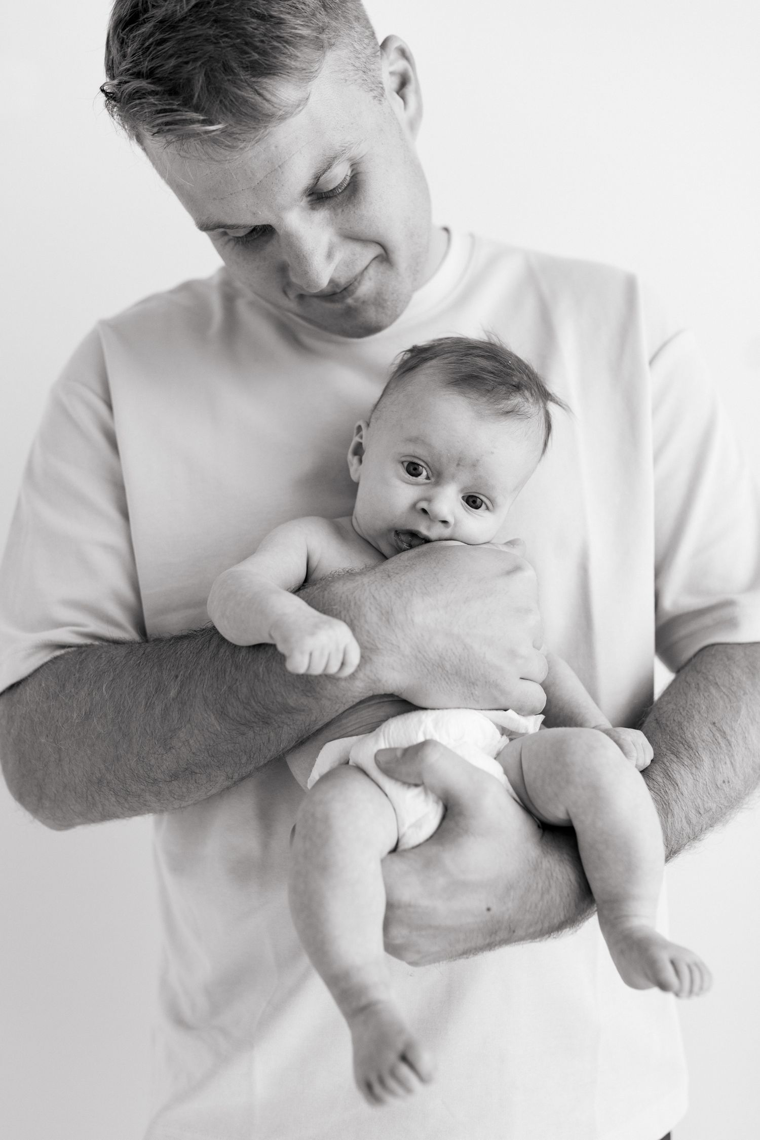 Tuore isä valkoisessa paidassa, sylissä poikavauva pelkässä vaipassa. Mustavalkoinen newborn-kuva studiolta.