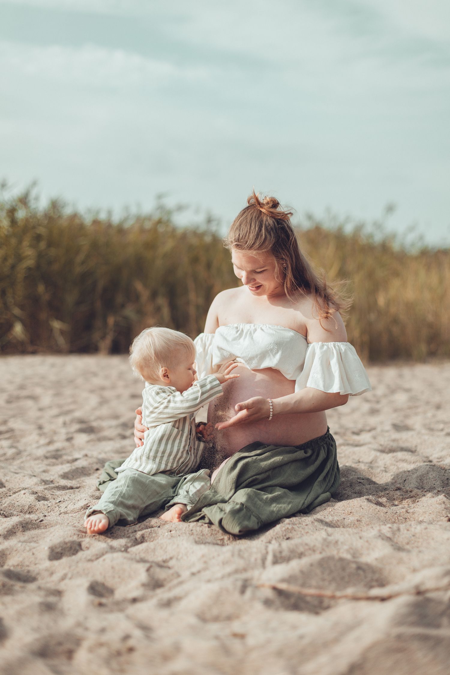 Pregnant woman in white top sitting on sandy beach with curious toddler touching her belly. Odotuskuvaus perheen kera miljöössä.