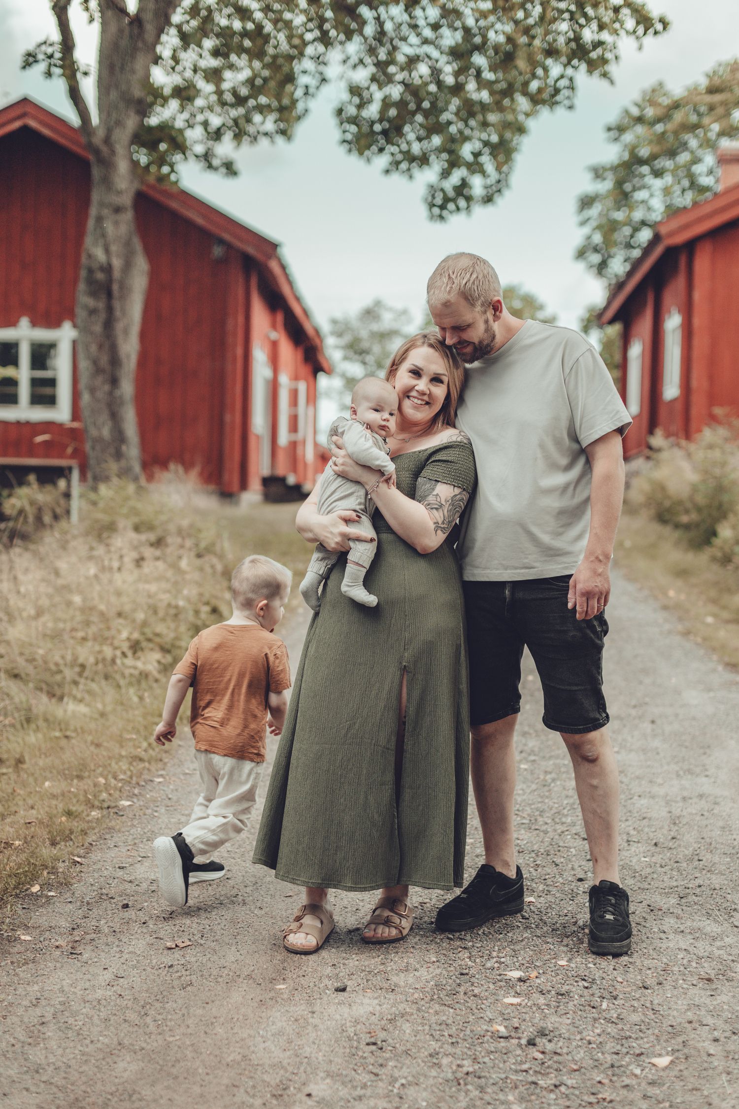Happy young family with baby and toddler standing on rural path beside red Scandinavian buildings. Perhekuvaus Fagervik.