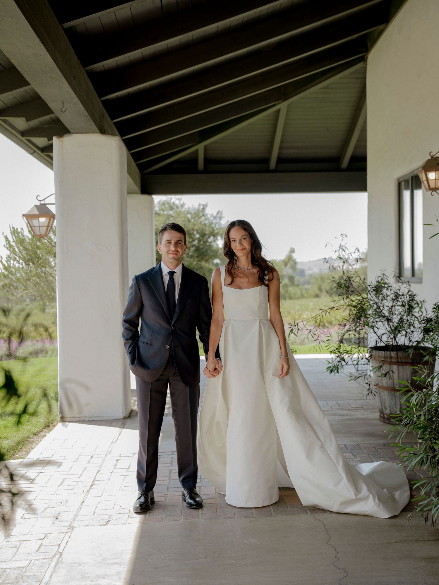 A couple in formal wedding attire poses under a covered walkway with white columns.