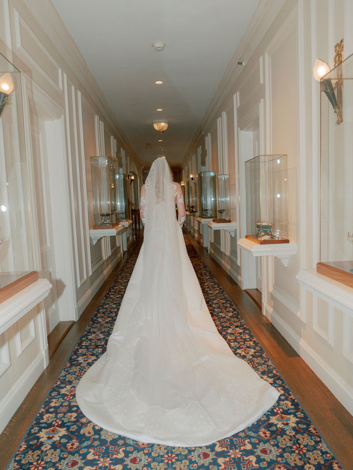 Long white wedding dress train flowing down an elegant hotel hallway with patterned carpet.