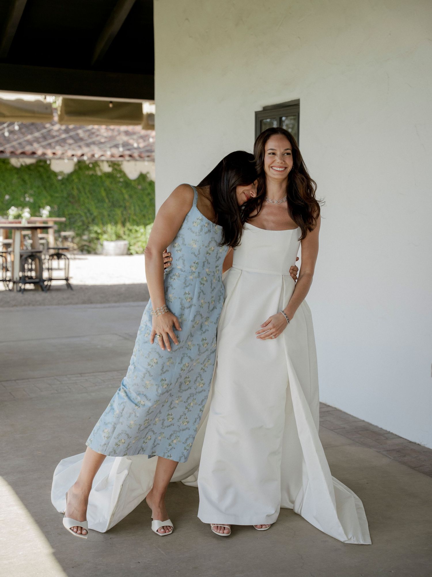 Two women in wedding attire sharing a candid moment on a covered porch.