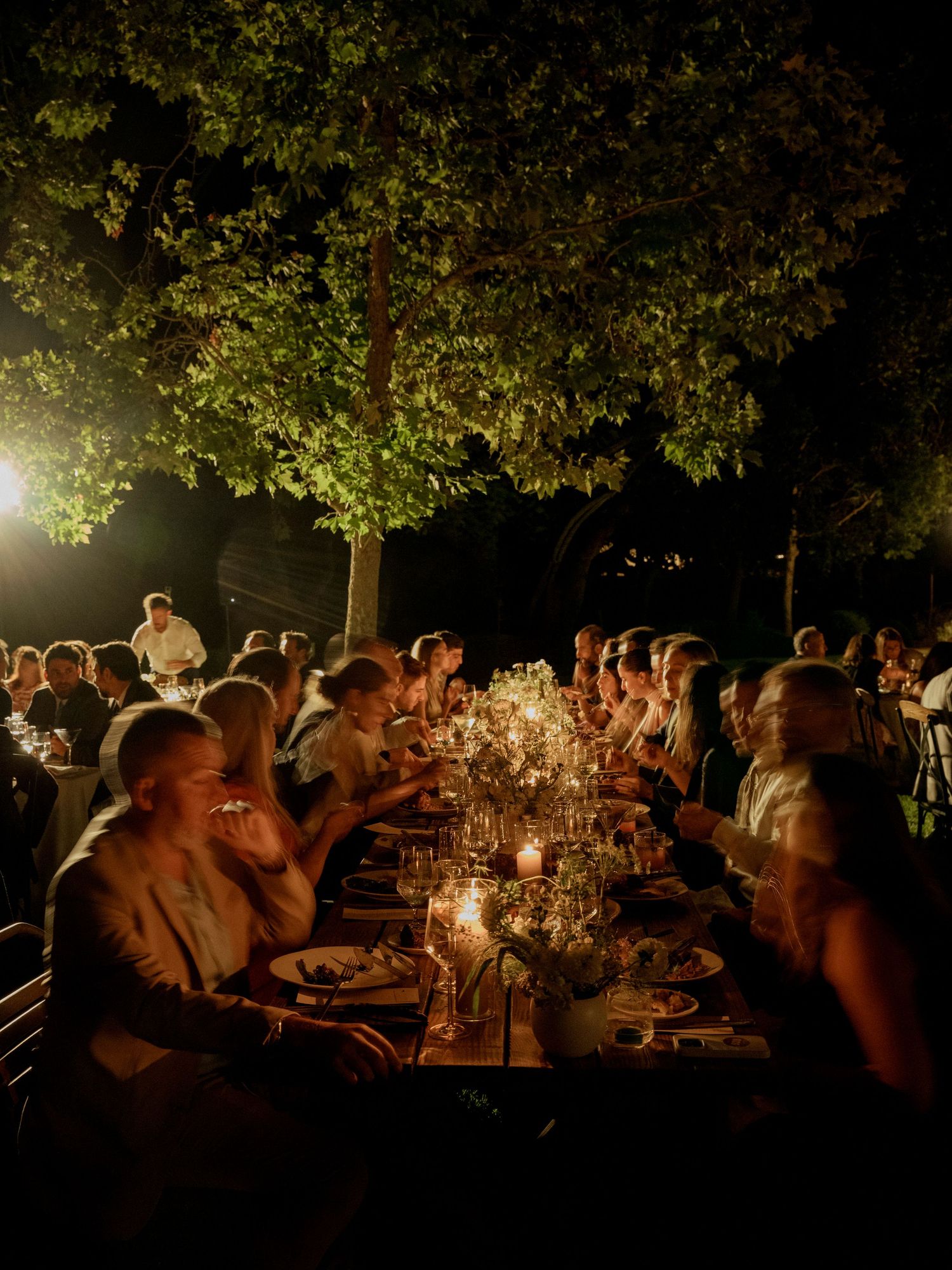 Wedding guests enjoy an intimate outdoor dinner reception under trees lit by warm candlelight.