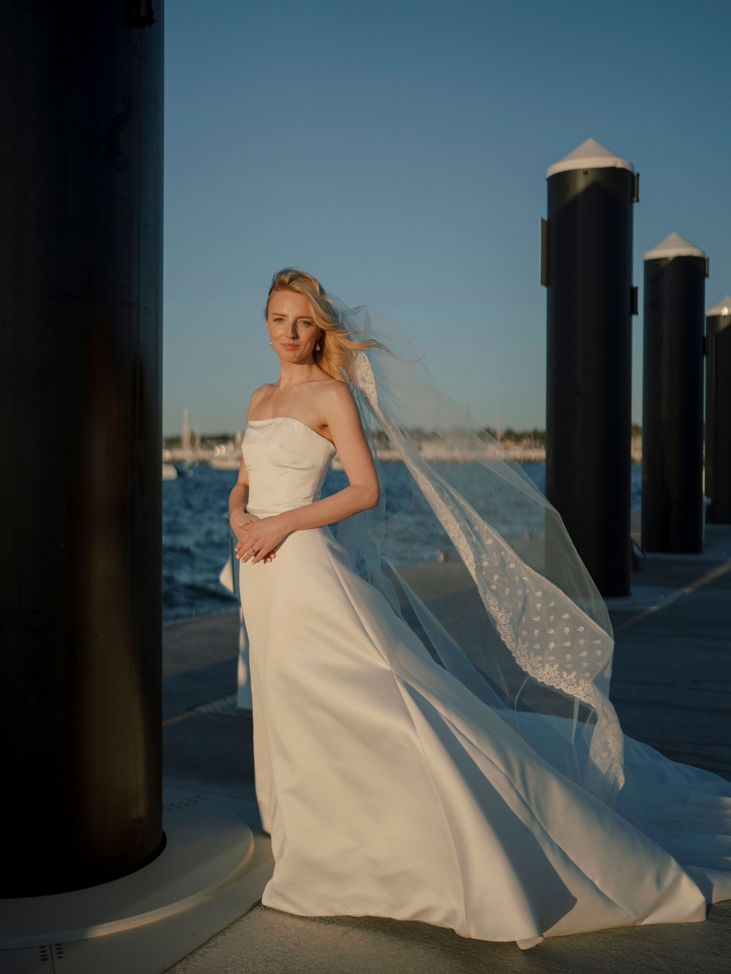 A bride in a flowing white dress poses against pillars near the waterfront at sunset.