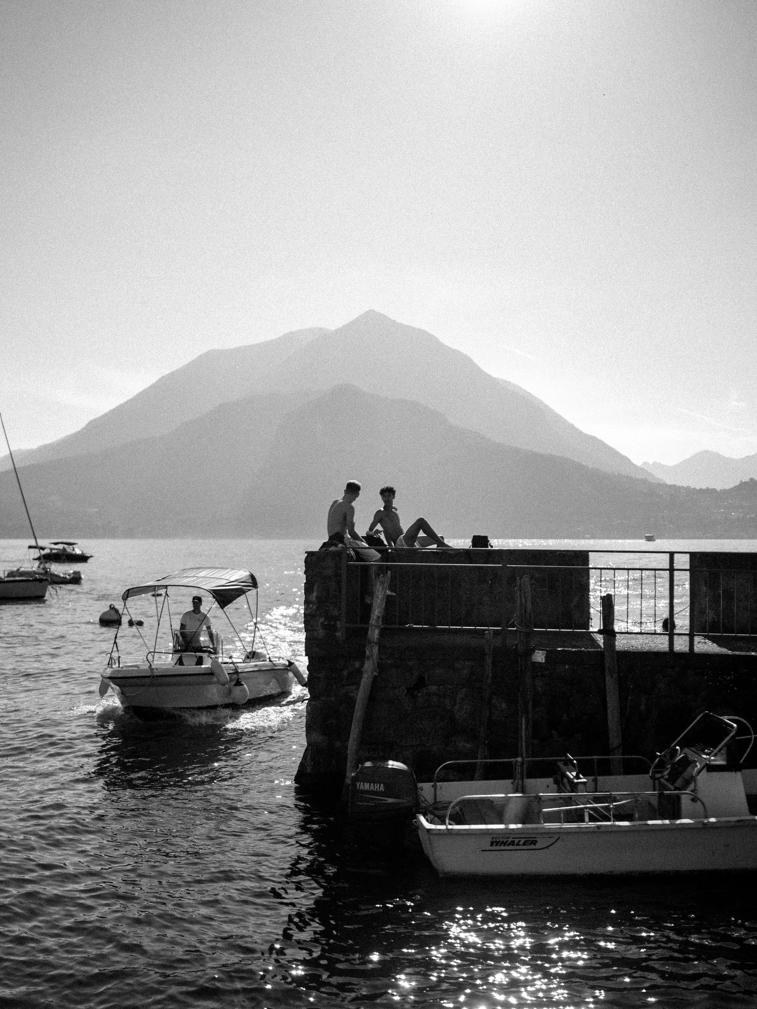 Black and white photograph of boats and people silhouetted against a mountainous lake view.