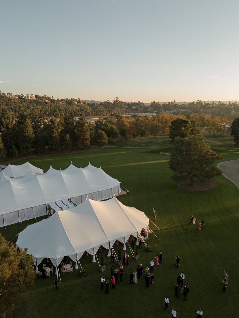 Large white wedding tent set up on a grassy lawn during golden hour with guests gathering outside.