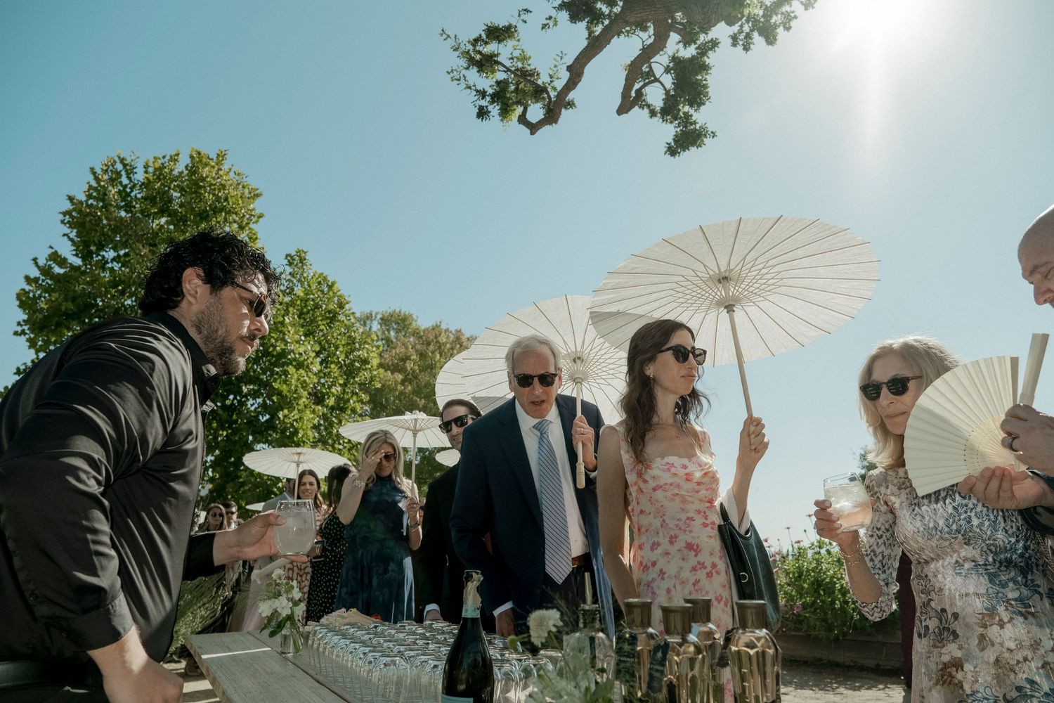 Wedding guests hold white parasols during an outdoor ceremony on a sunny day.