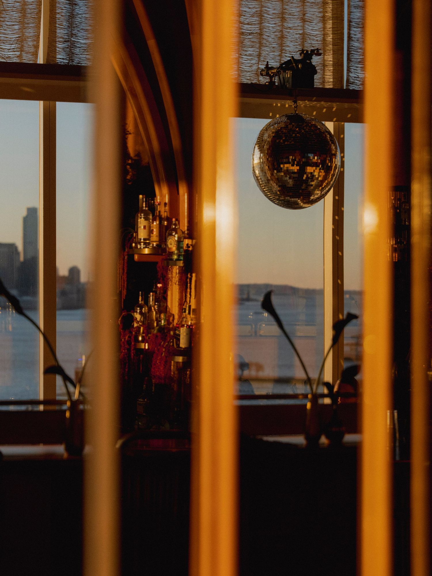 A disco ball hangs in a dimly lit venue with city views through large windows at sunset.