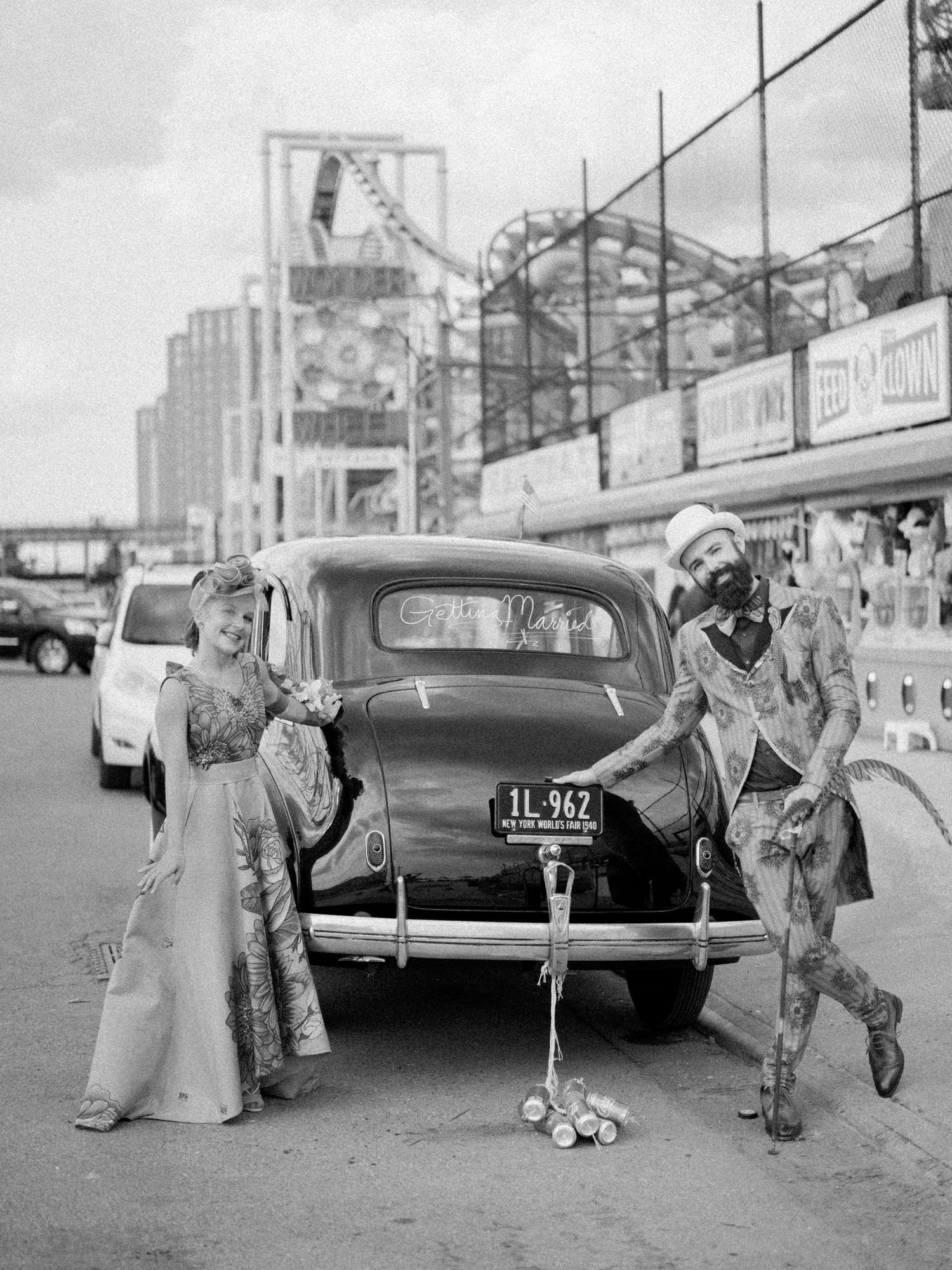 Vintage black and white photo of performers in costume on a boardwalk near classic cars.