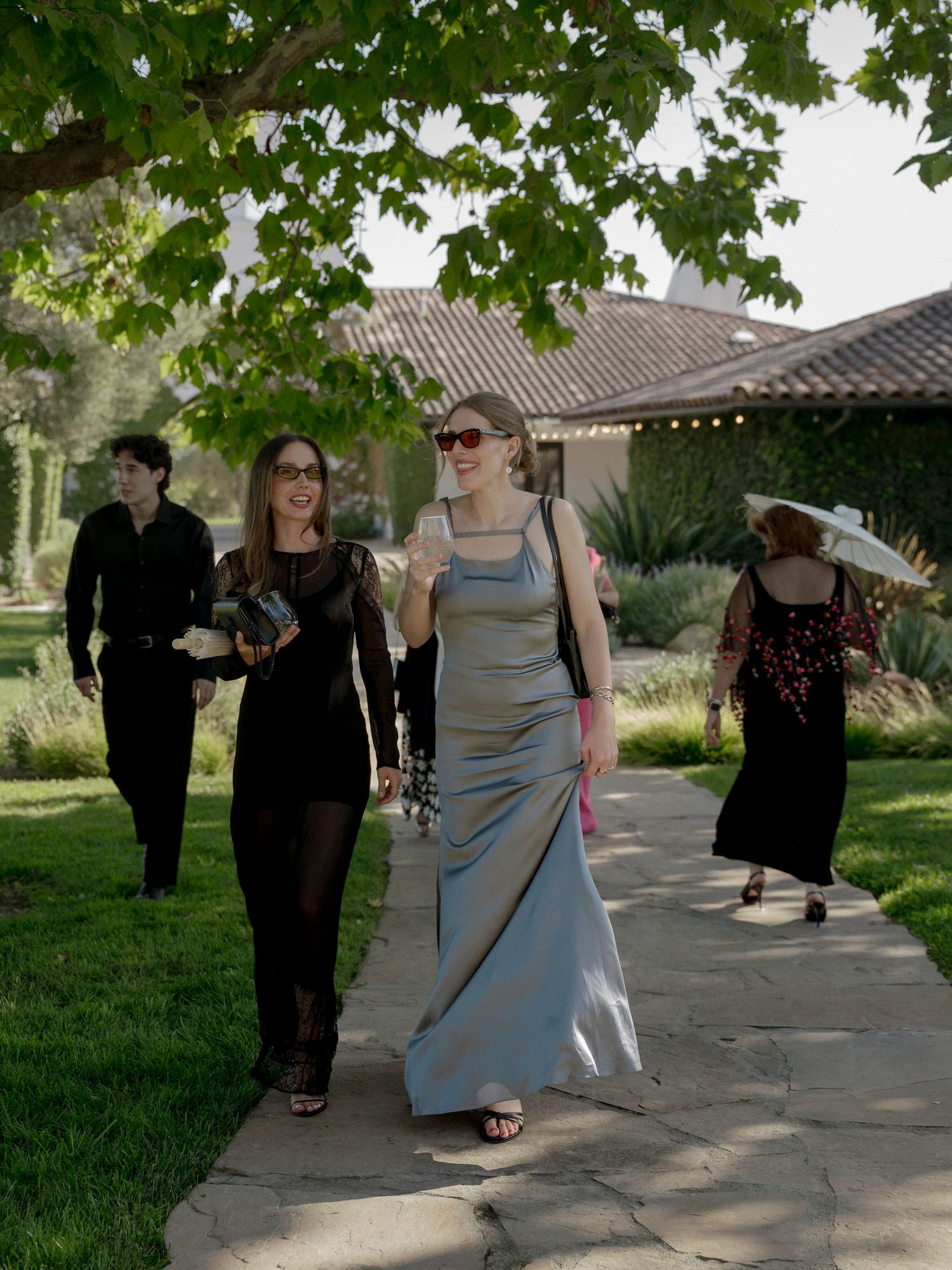 Wedding guests walking along a stone pathway under green trees.