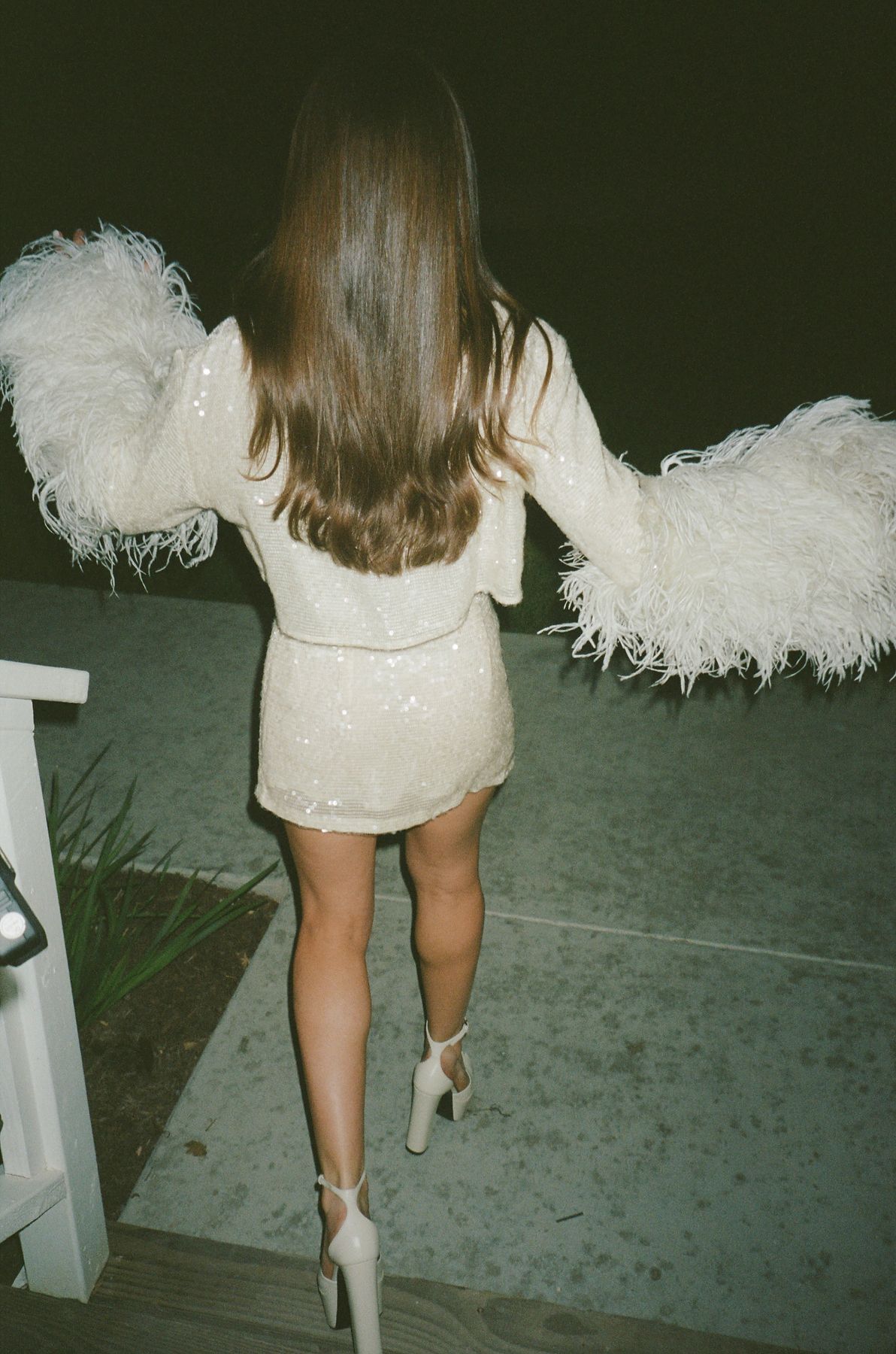 Close-up view of a white feathered dress hem and heels walking on a dark surface.
