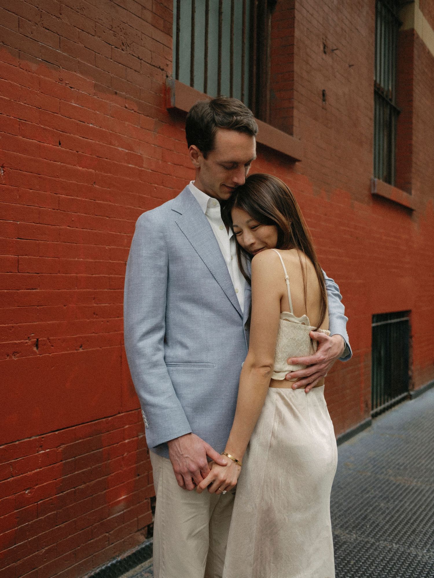 Couple in formal attire embracing against a rustic red brick wall.