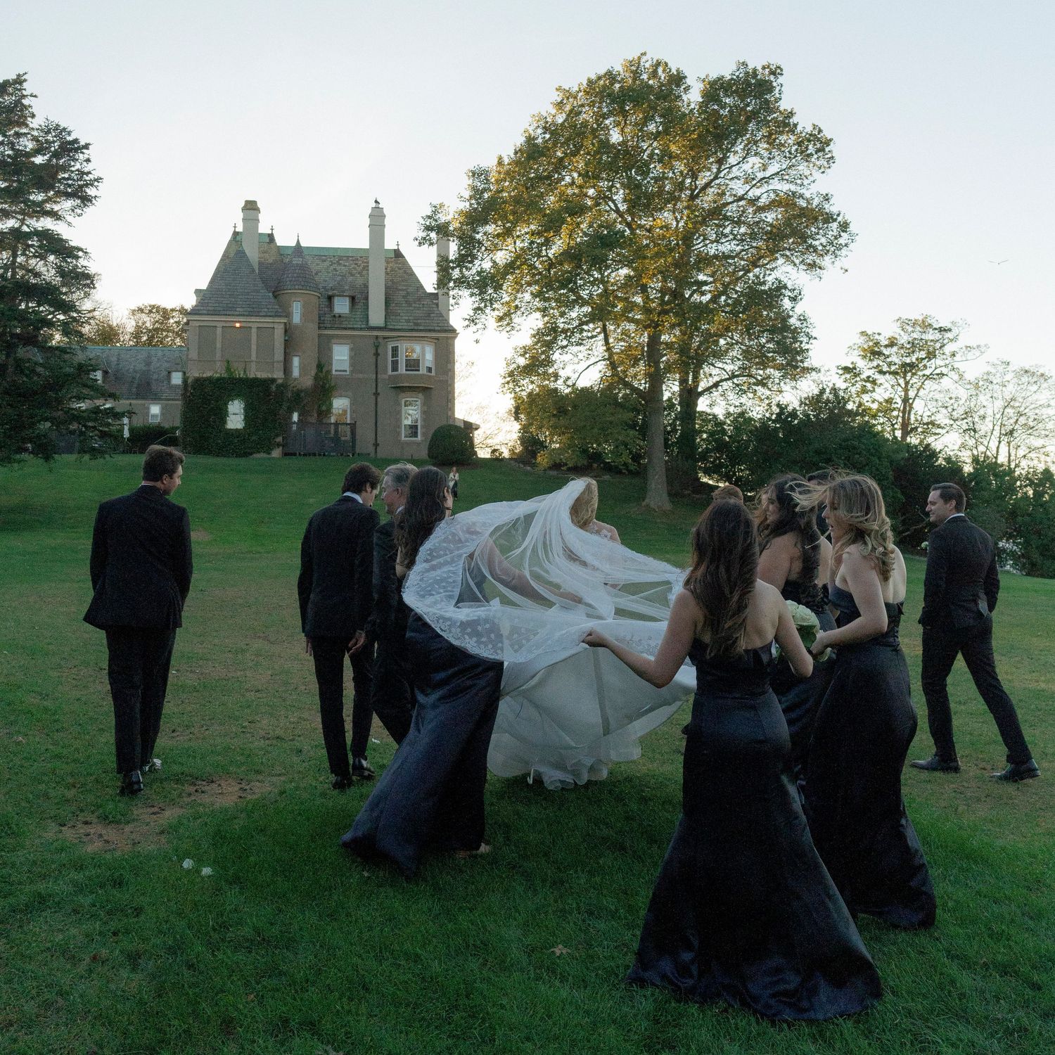 Wedding party members dance and celebrate on a grassy lawn with a historic building in the background.