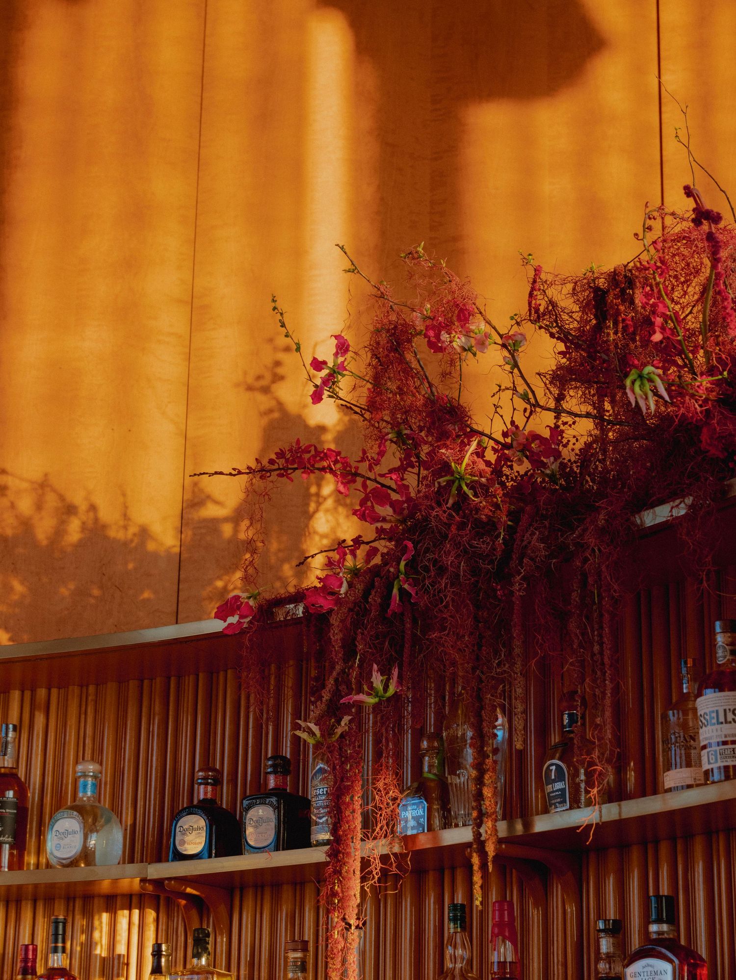Decorative red floral arrangements adorn a wooden shelf displaying various liquor bottles in warm lighting.