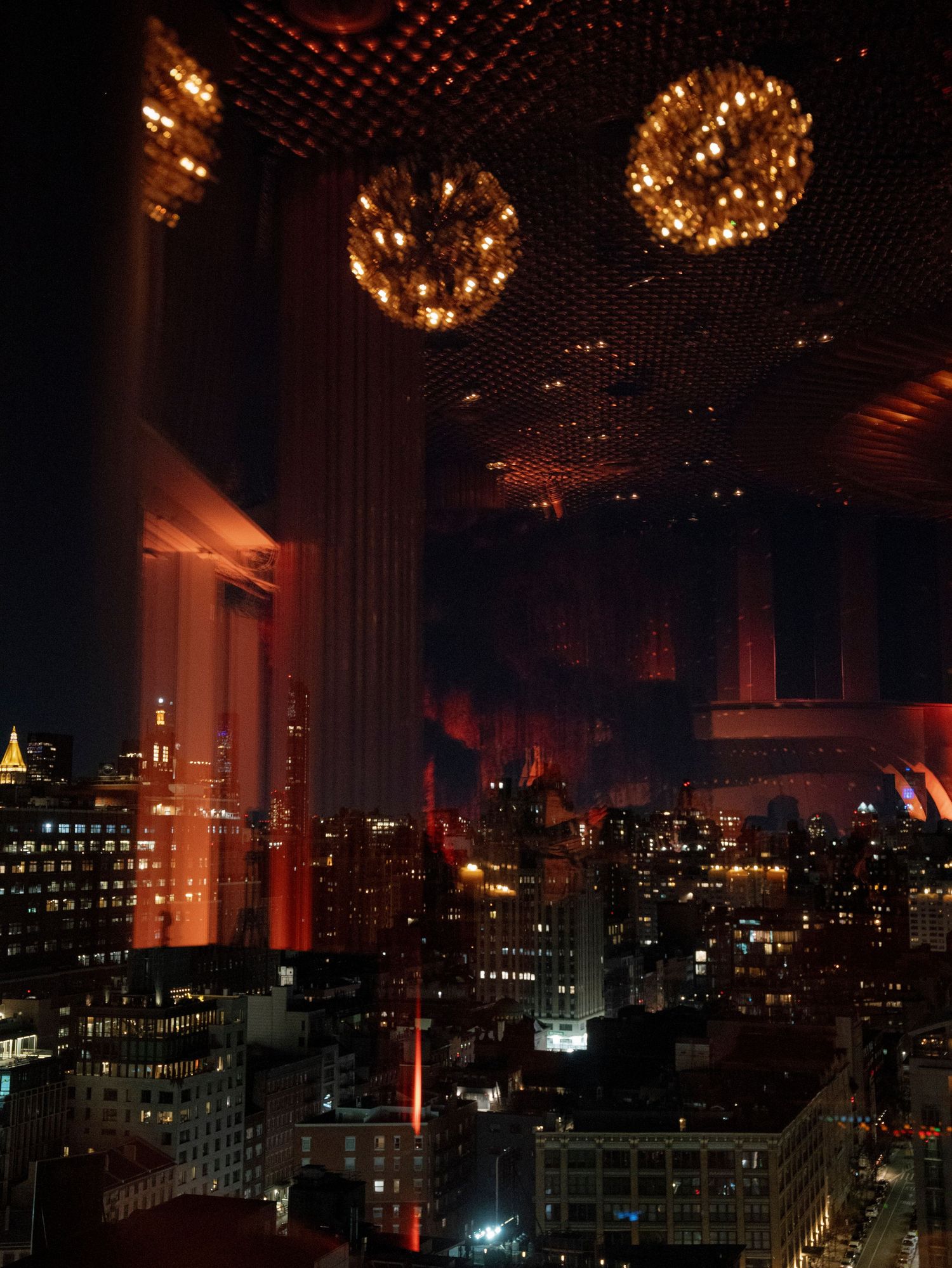 Illuminated spherical chandeliers hang above a cityscape view through large windows at night.