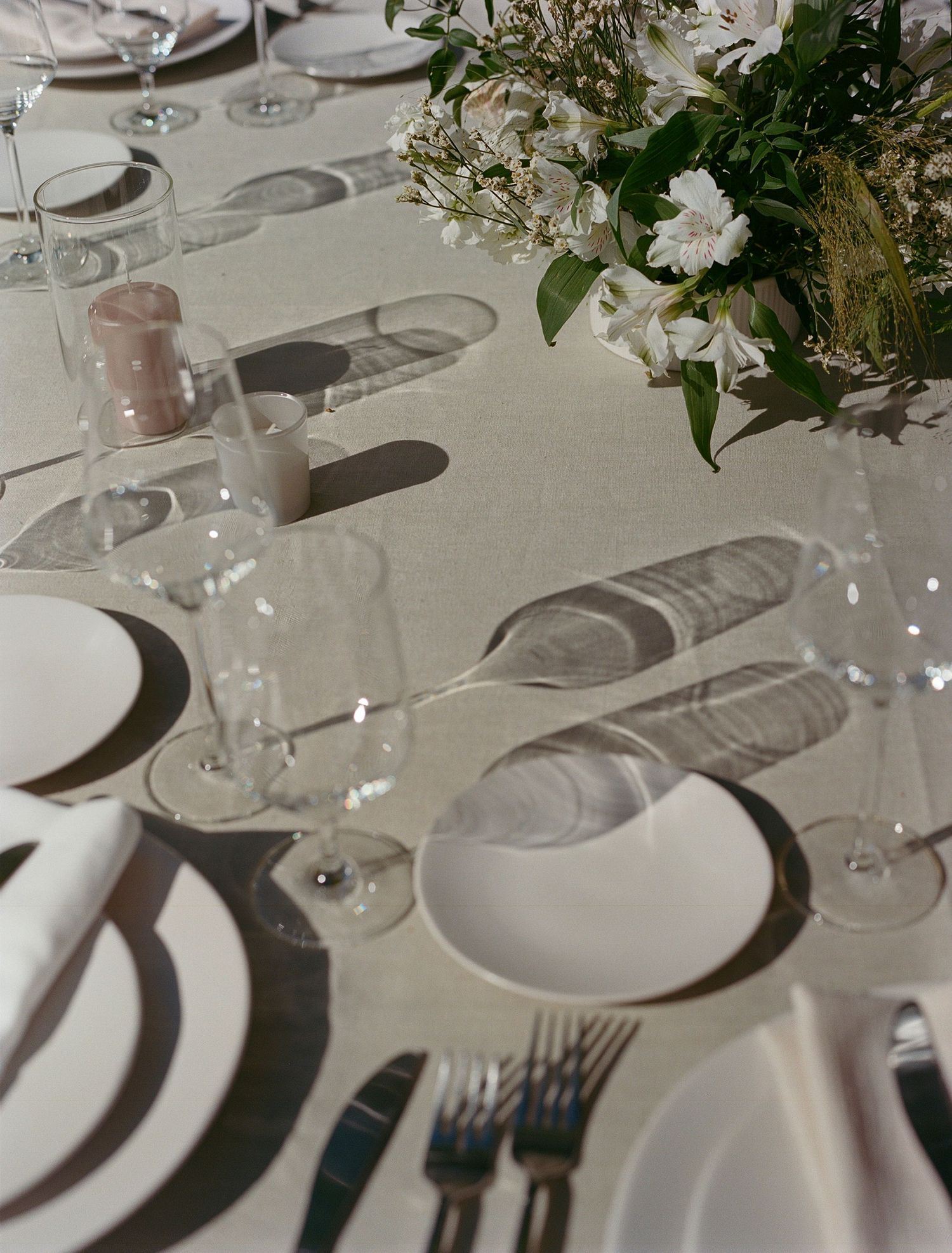 Elegant table setting with white plates, wine glasses and floral arrangements casting soft shadows.