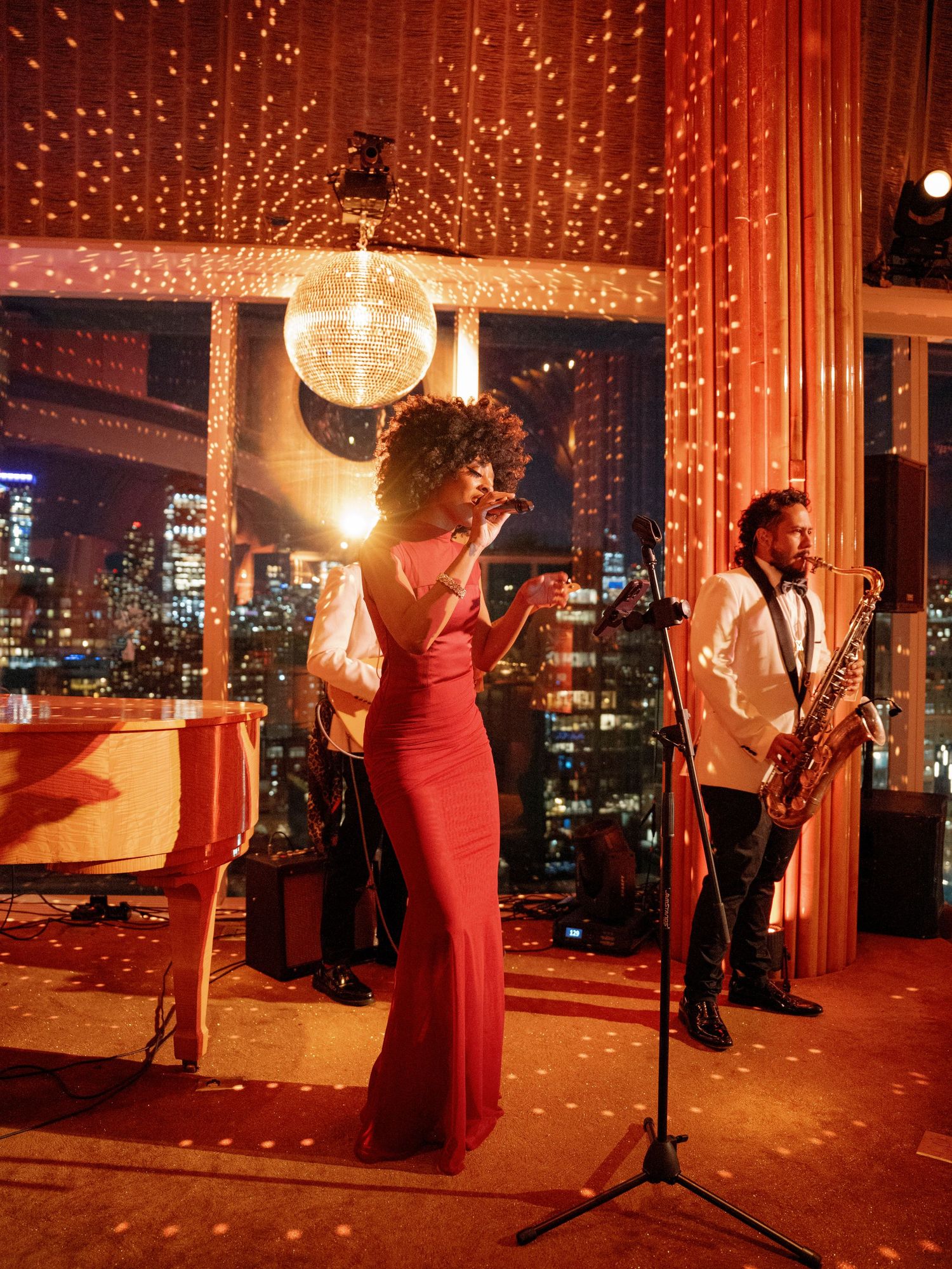 A live jazz performance featuring musicians and a singer in a red dress against a city skyline backdrop with twinkling lights.