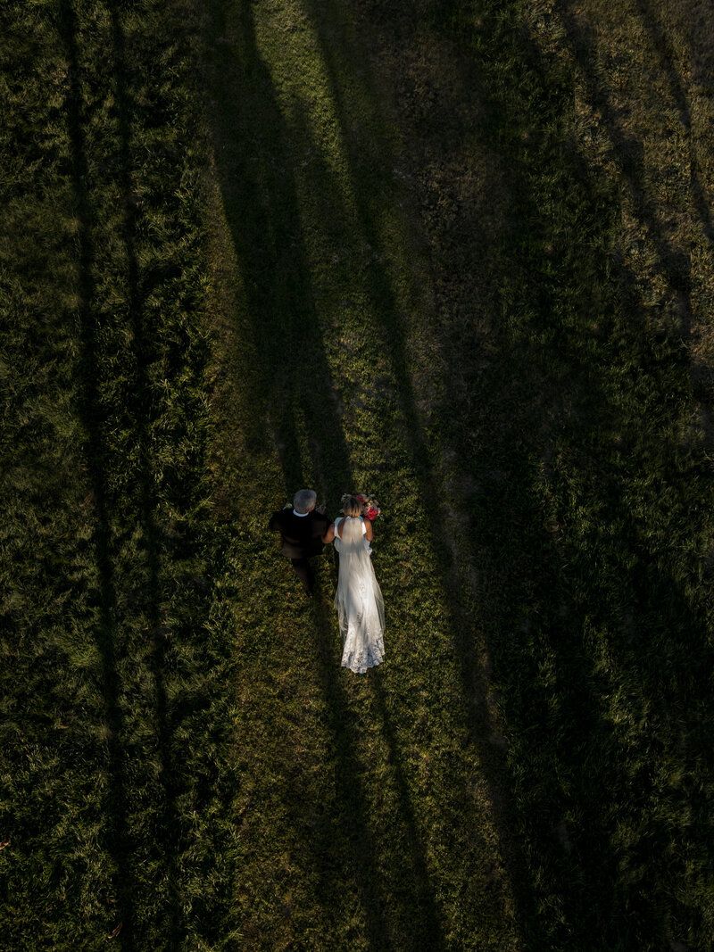 Aerial view of couple walking through a green field with dramatic shadows from trees.