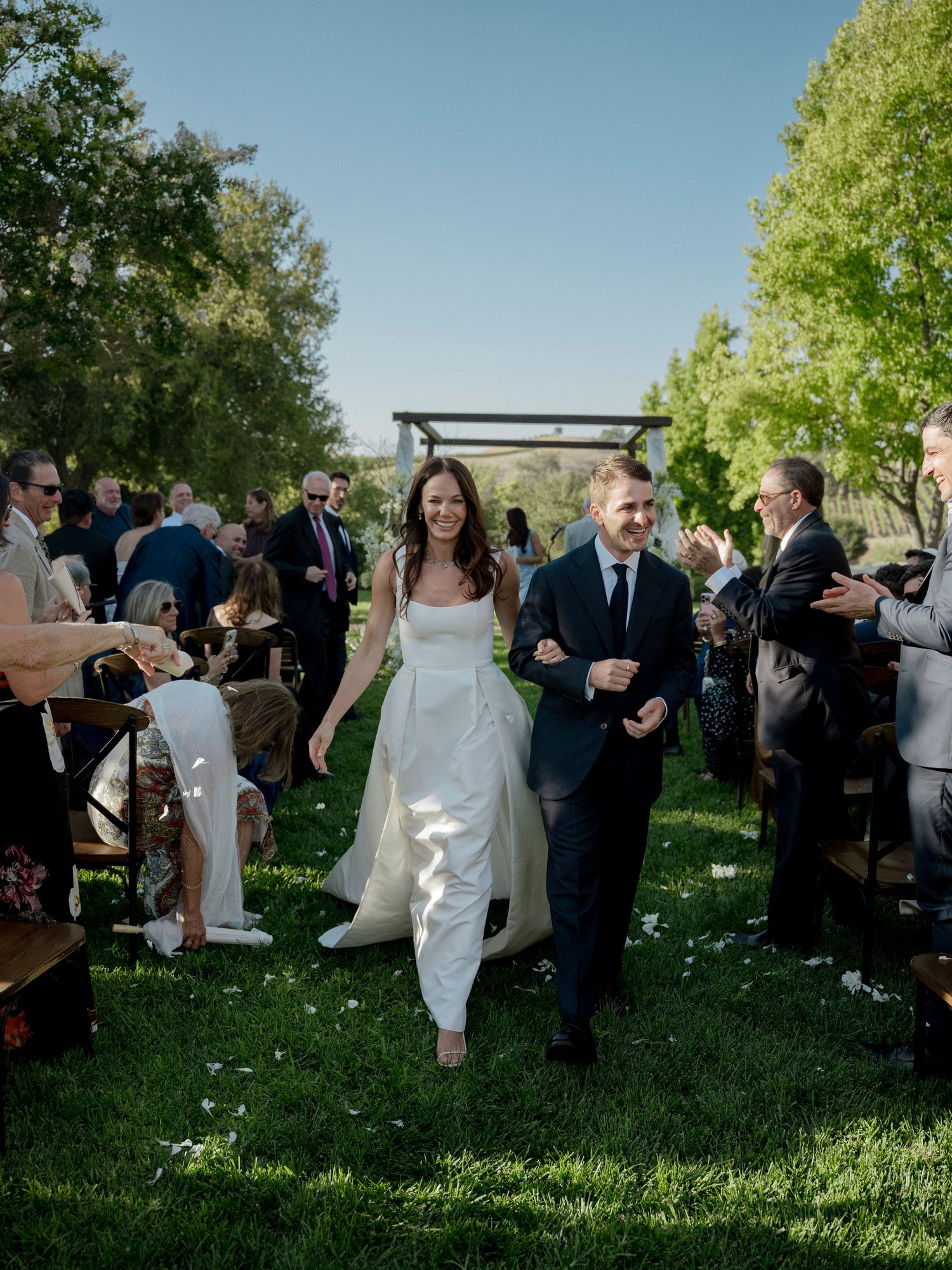 Wedding couple walking down aisle while guests celebrate on lawn with wooden chairs.