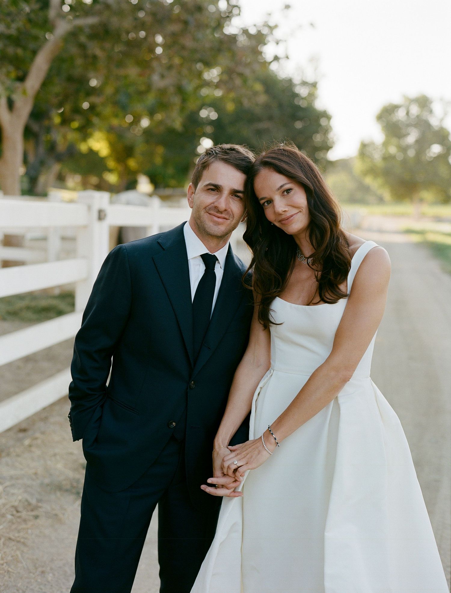 Newlyweds sharing intimate moment while posing by white fence on country road.