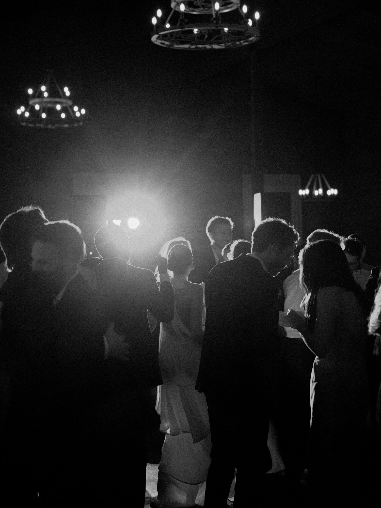 Silhouettes of party guests dancing under dimly lit chandeliers at a reception.