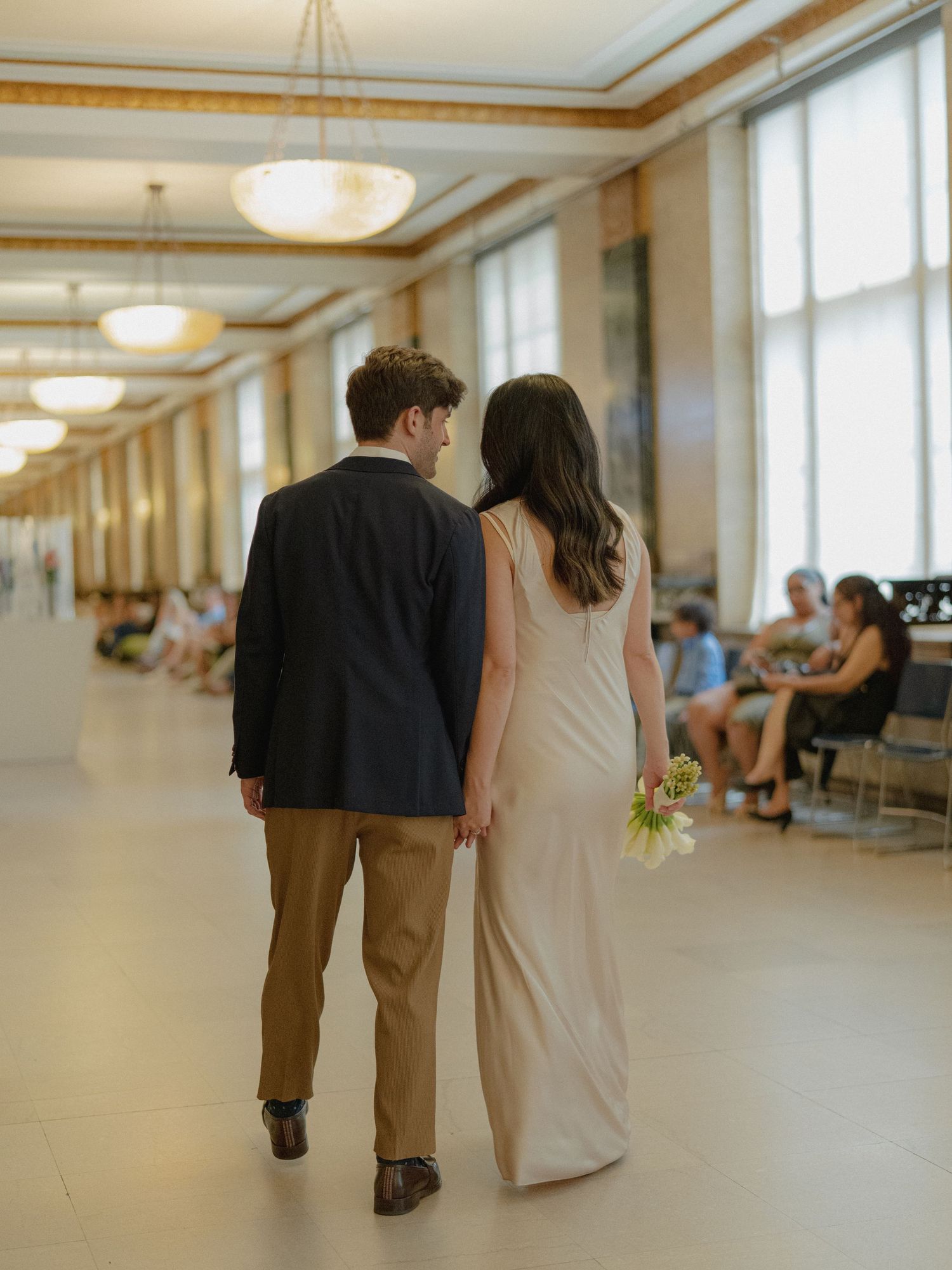 A couple walks down a sunlit hallway with elegant chandeliers and large windows.
