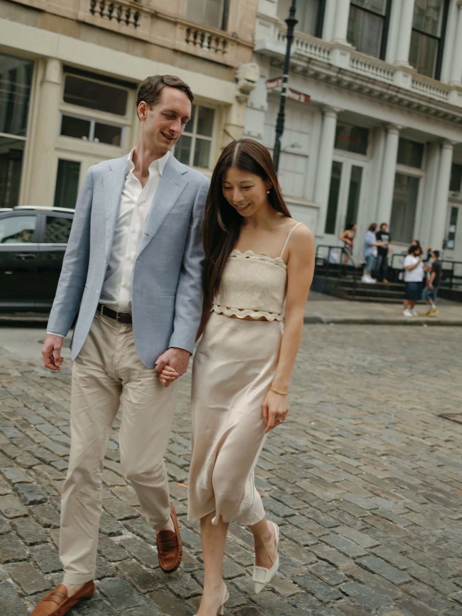 Couple walking playfully on cobblestone street in coordinated formal outfits.