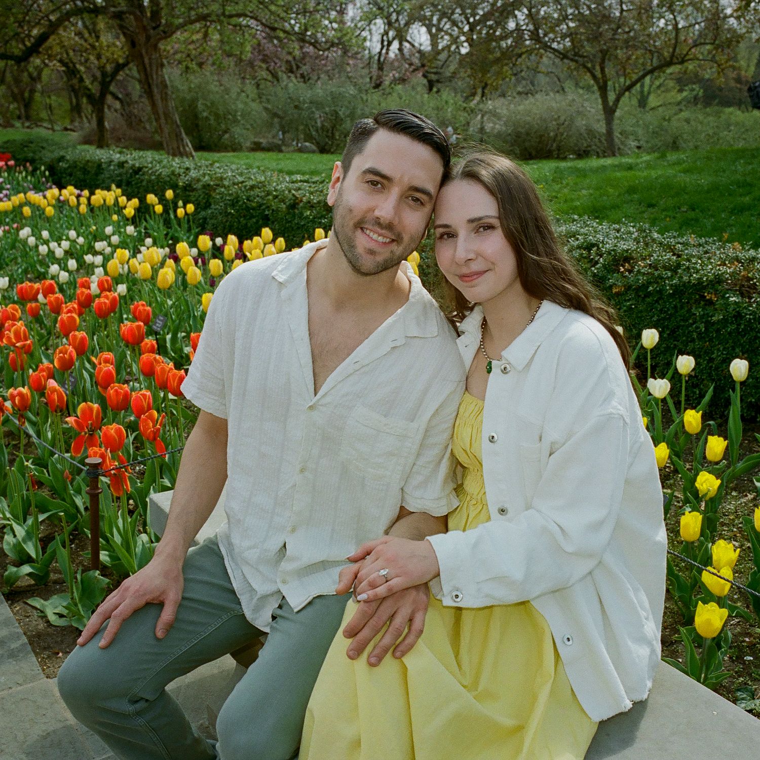 A couple poses among colorful spring tulips in a garden setting wearing white and yellow outfits.