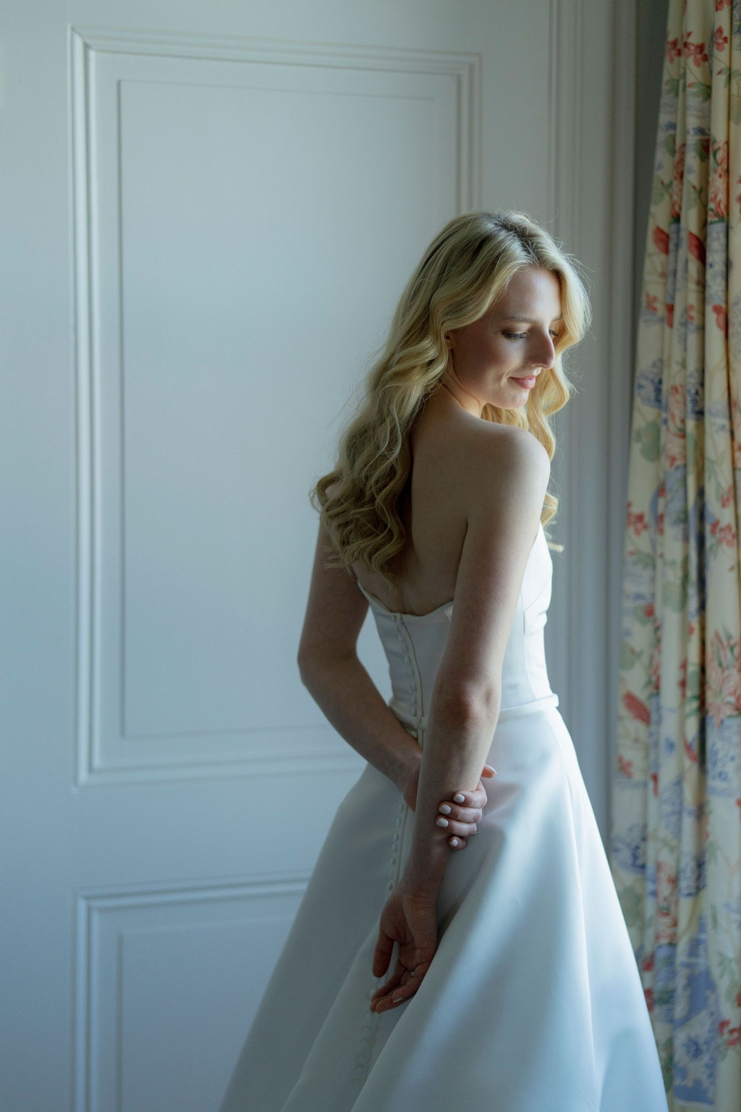 Bride in elegant white wedding gown standing in sunlit room with classic decor.