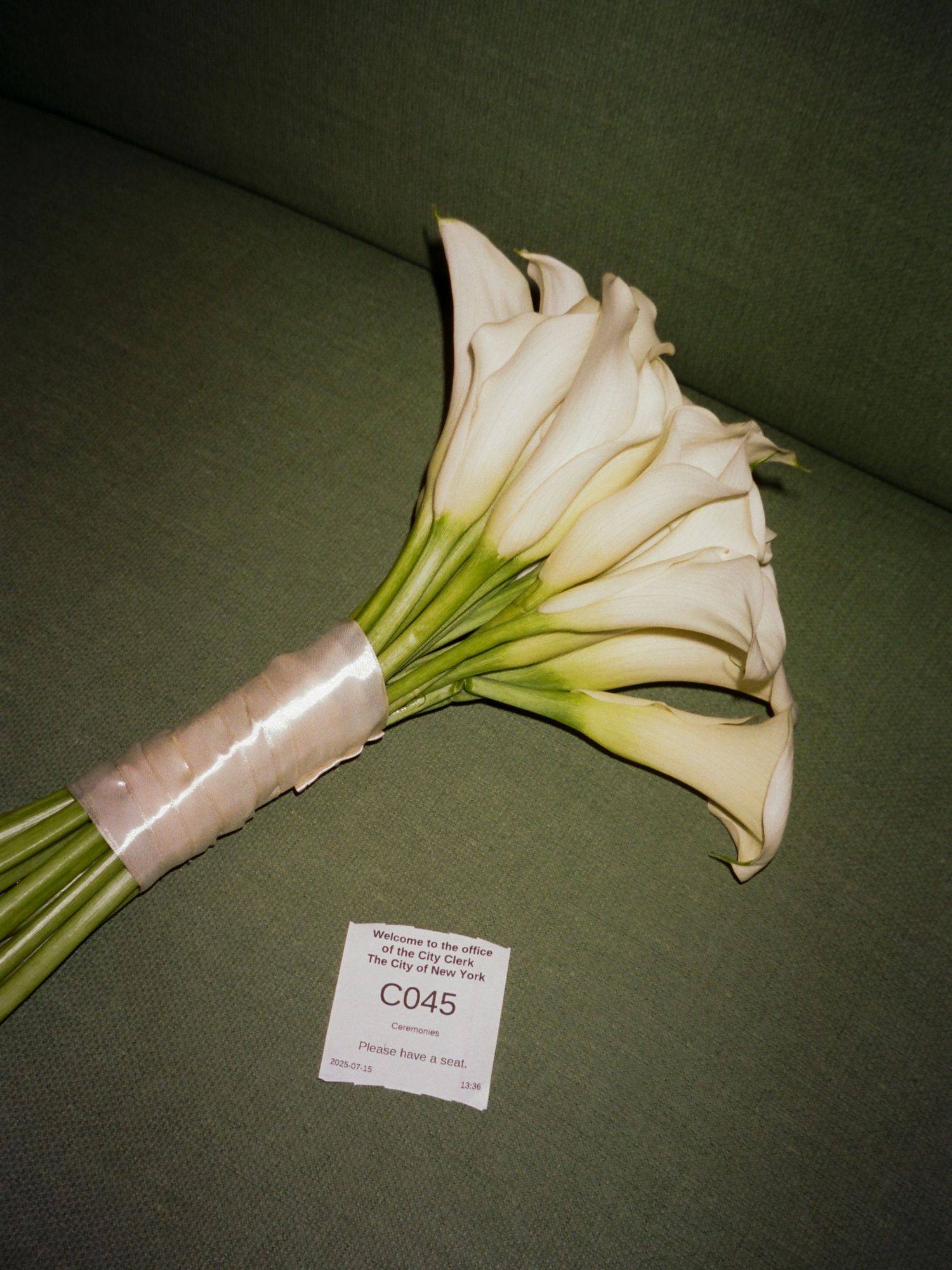 A elegant white calla lily bouquet wrapped with a blush pink ribbon rests on a dark green surface.