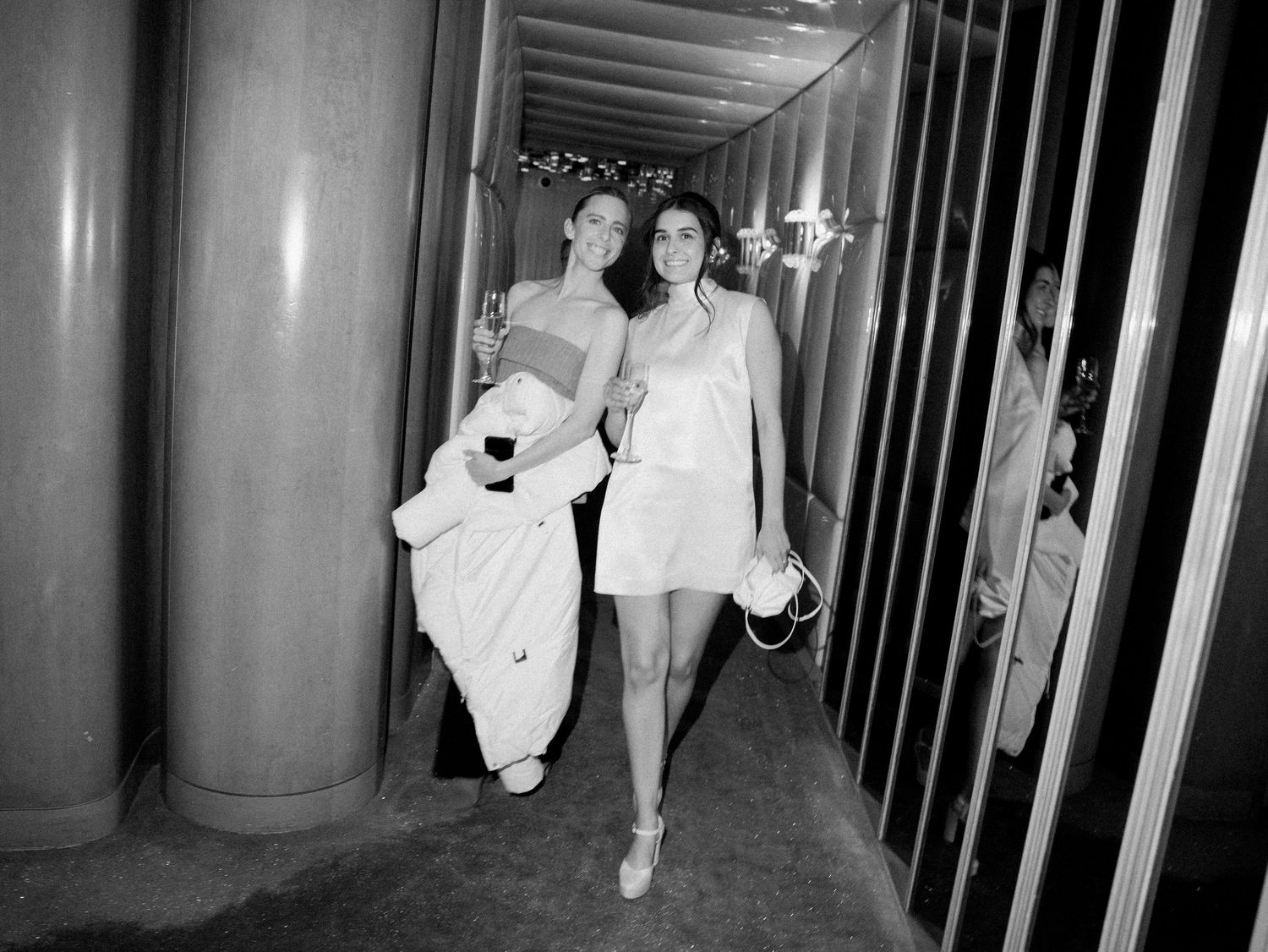 Two celebrants walking through a modern hallway in evening attire.