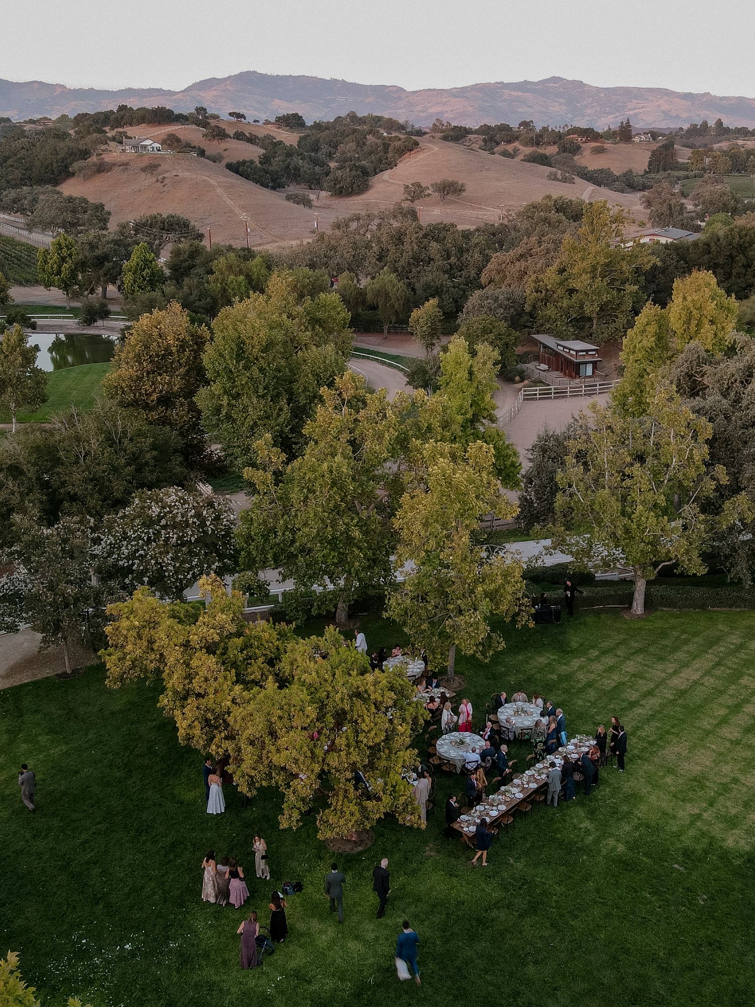 Aerial view of an outdoor wedding reception setup with tables arranged on a lush green lawn surrounded by autumn trees.