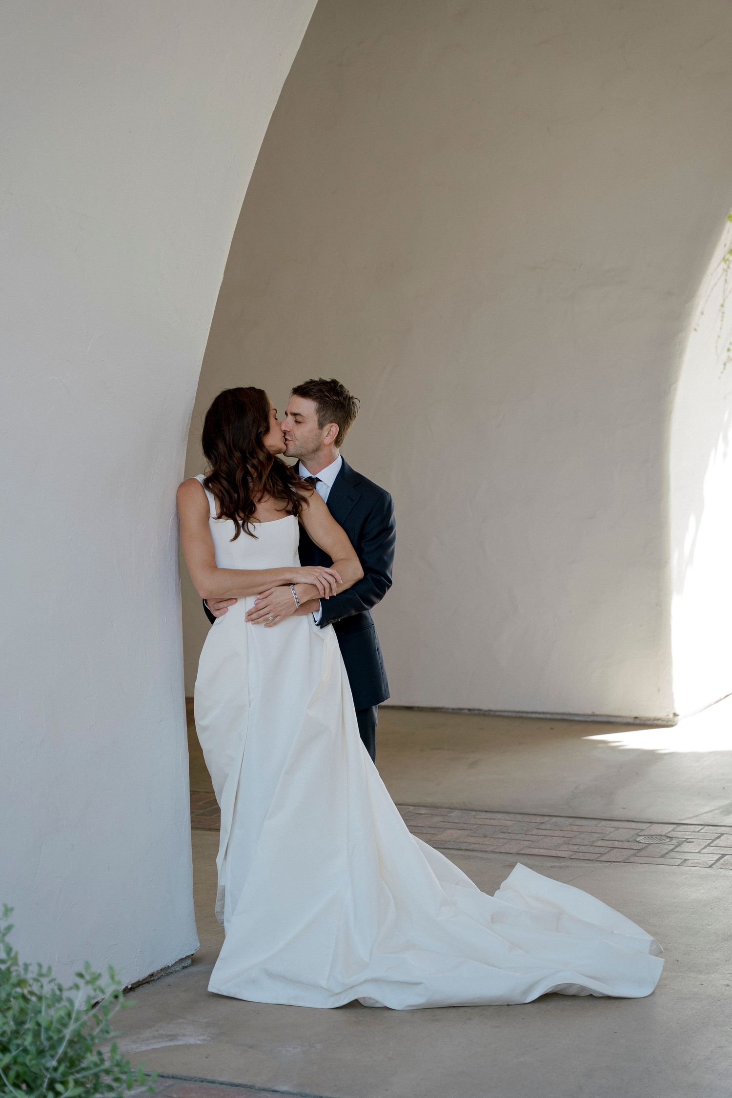A couple shares a romantic moment in white wedding attire against a light colored wall.