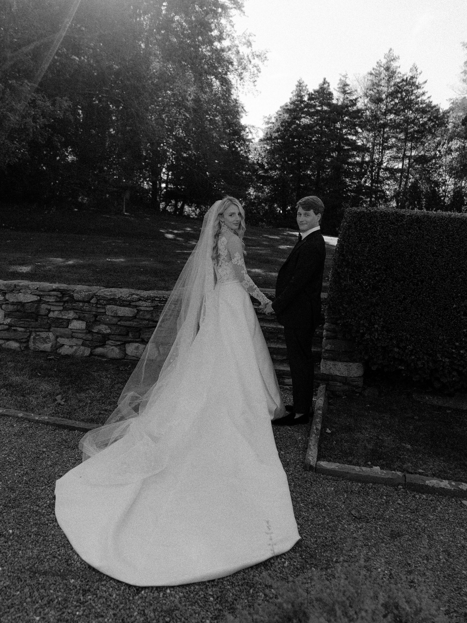 Black and white outdoor wedding photo showing a flowing white dress train against a stone wall backdrop.