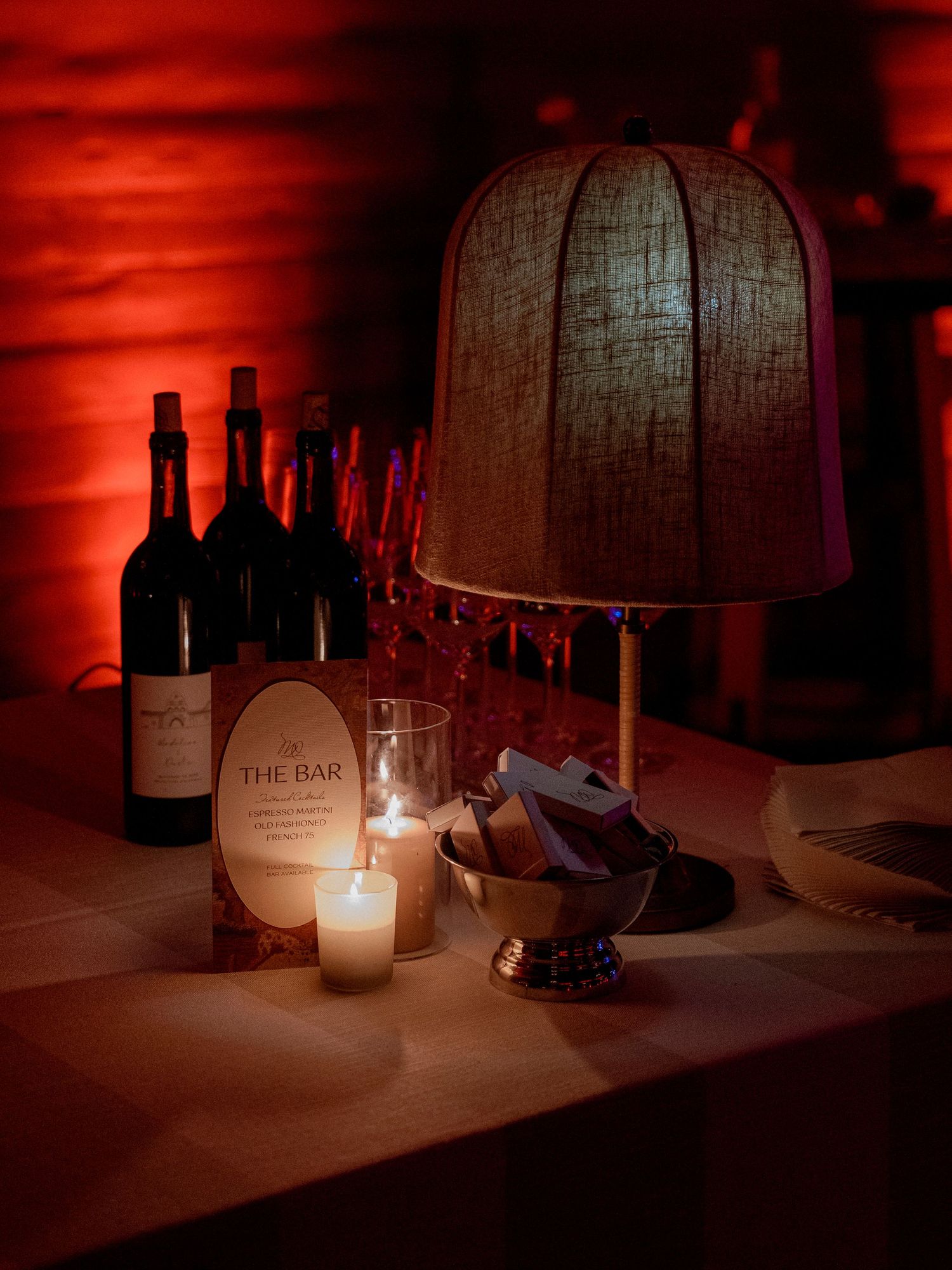 A moody bar setup with wine bottles and candles creating atmospheric red lighting.