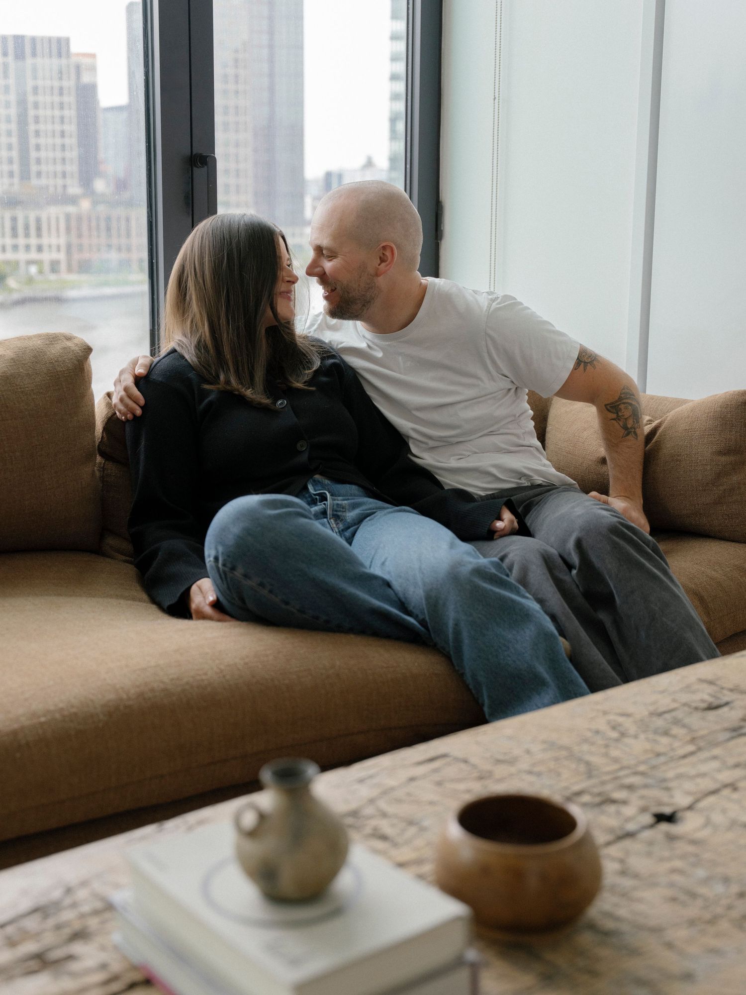 Couple relaxing together on a brown couch with city views through windows.
