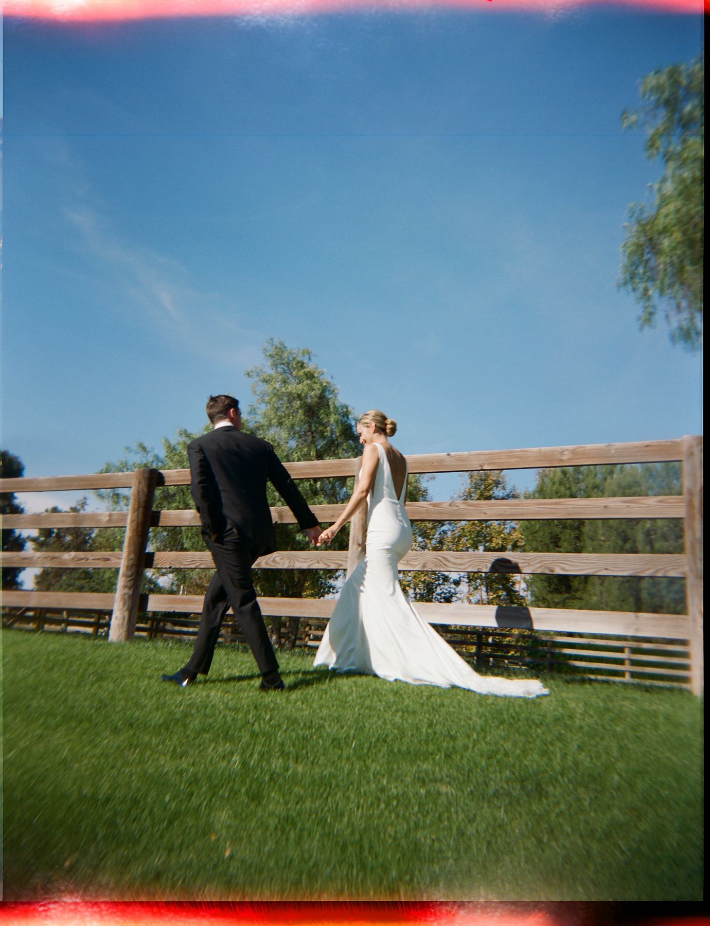Wedding couple sitting together on grass against a wooden fence at sunset.