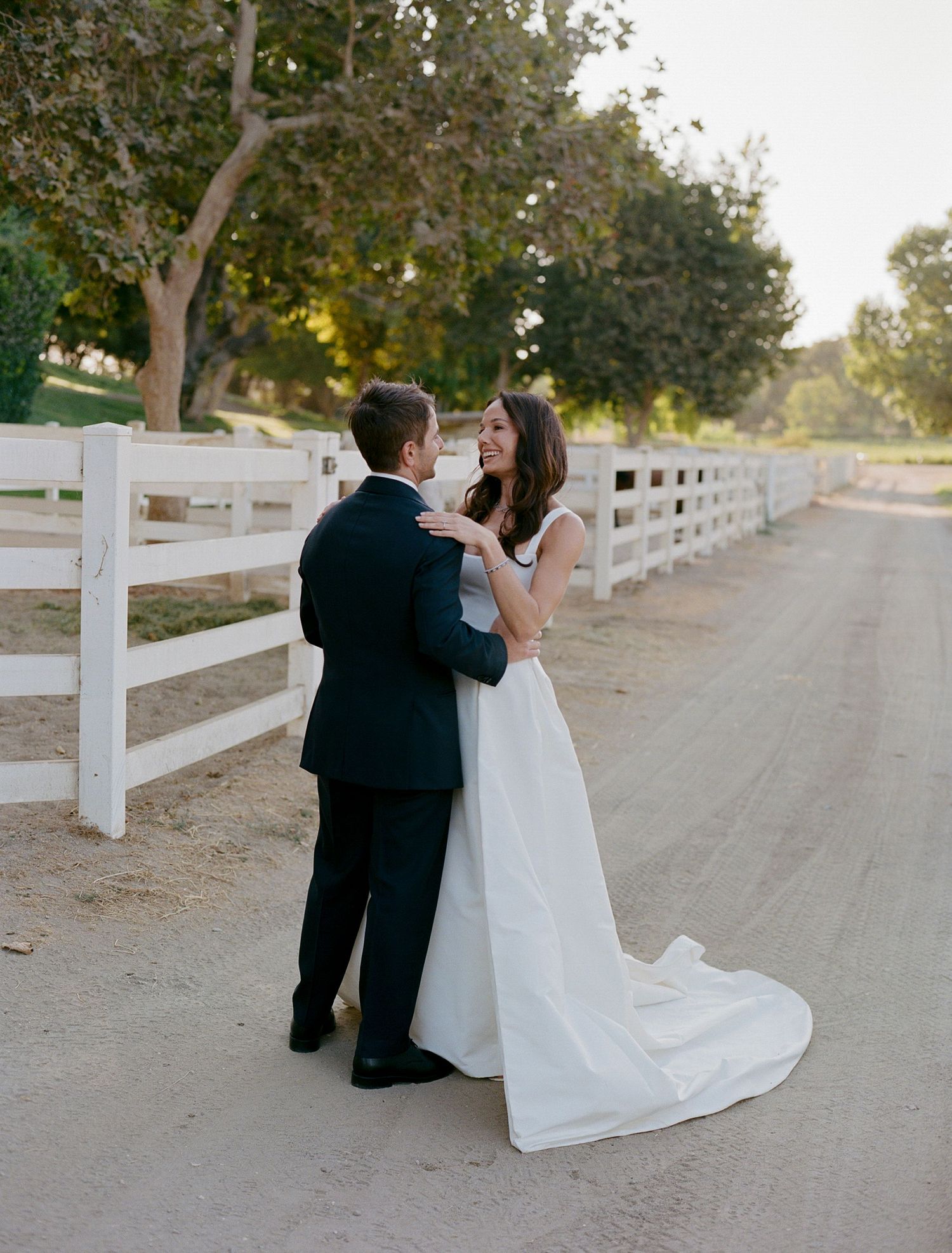 Couple sitting on a white fence-lined dirt path during a wedding photo session.