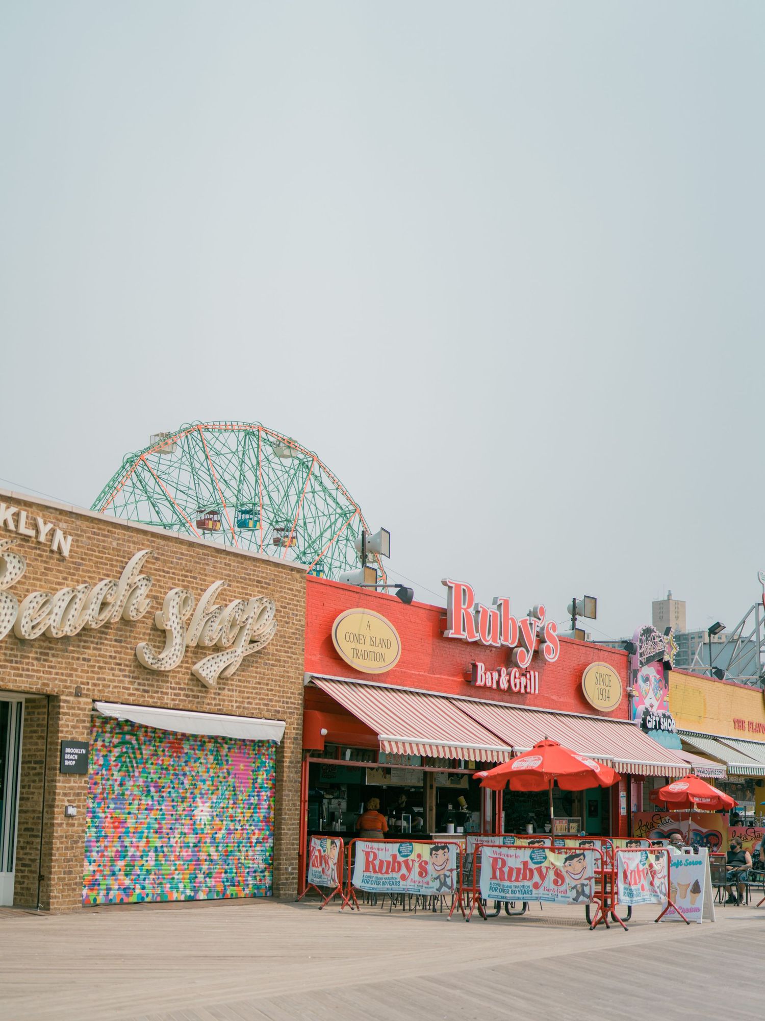 Vintage-style storefront panorama of beachside shops with colorful signage and a ferris wheel.