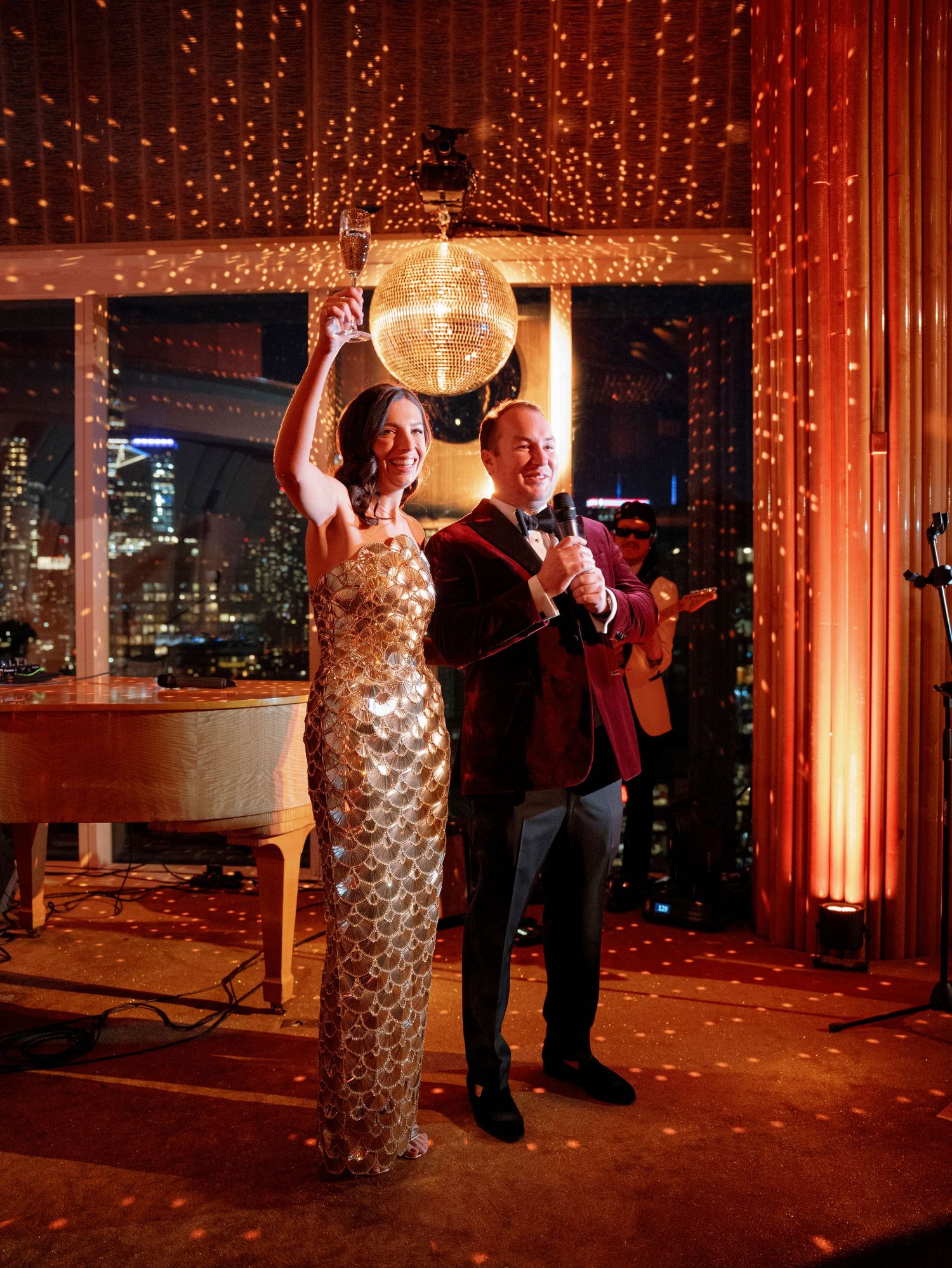 Couple dancing under a sparkling disco ball with city lights visible through windows in an elegant venue.