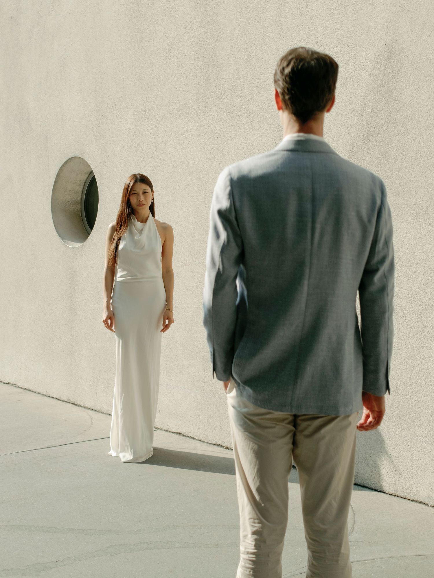 Two people in formal wear walking against a white wall with dramatic shadows.