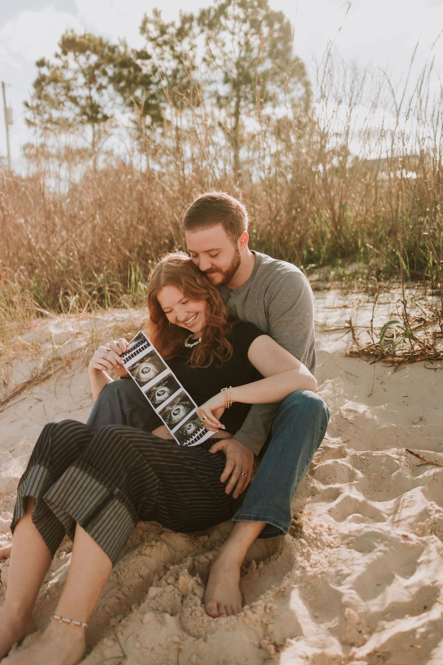 Couple sitting on beach sand holding ultrasound photos, announcing pregnancy outdoors near coastal dunes.