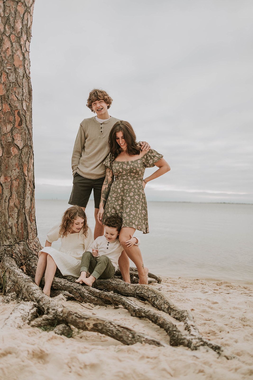A family enjoying a casual beach day together by a large tree trunk with an overcast sky in the background.