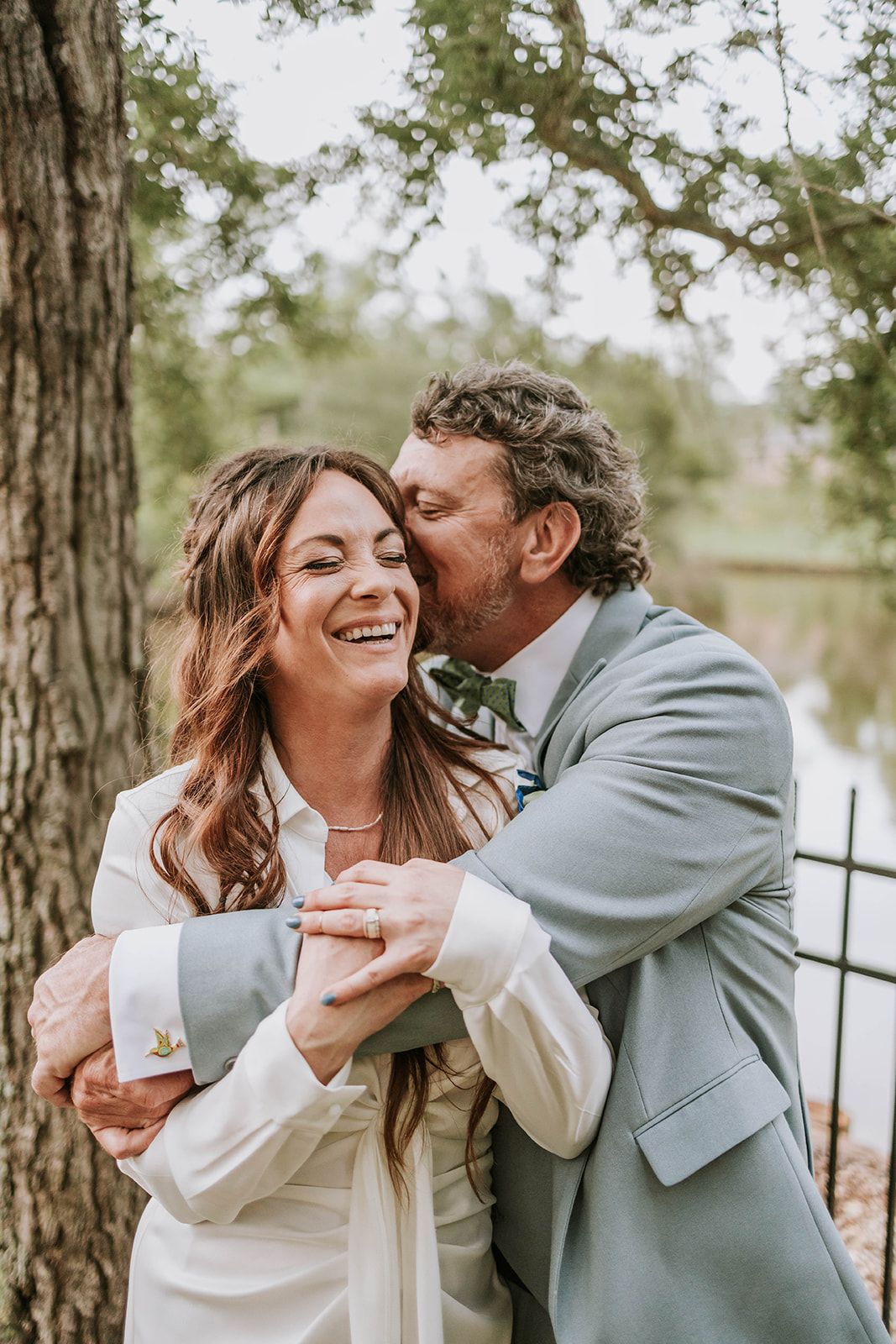 Romantic wedding couple embraces and laughs joyfully under a tree by the water in natural outdoor setting.