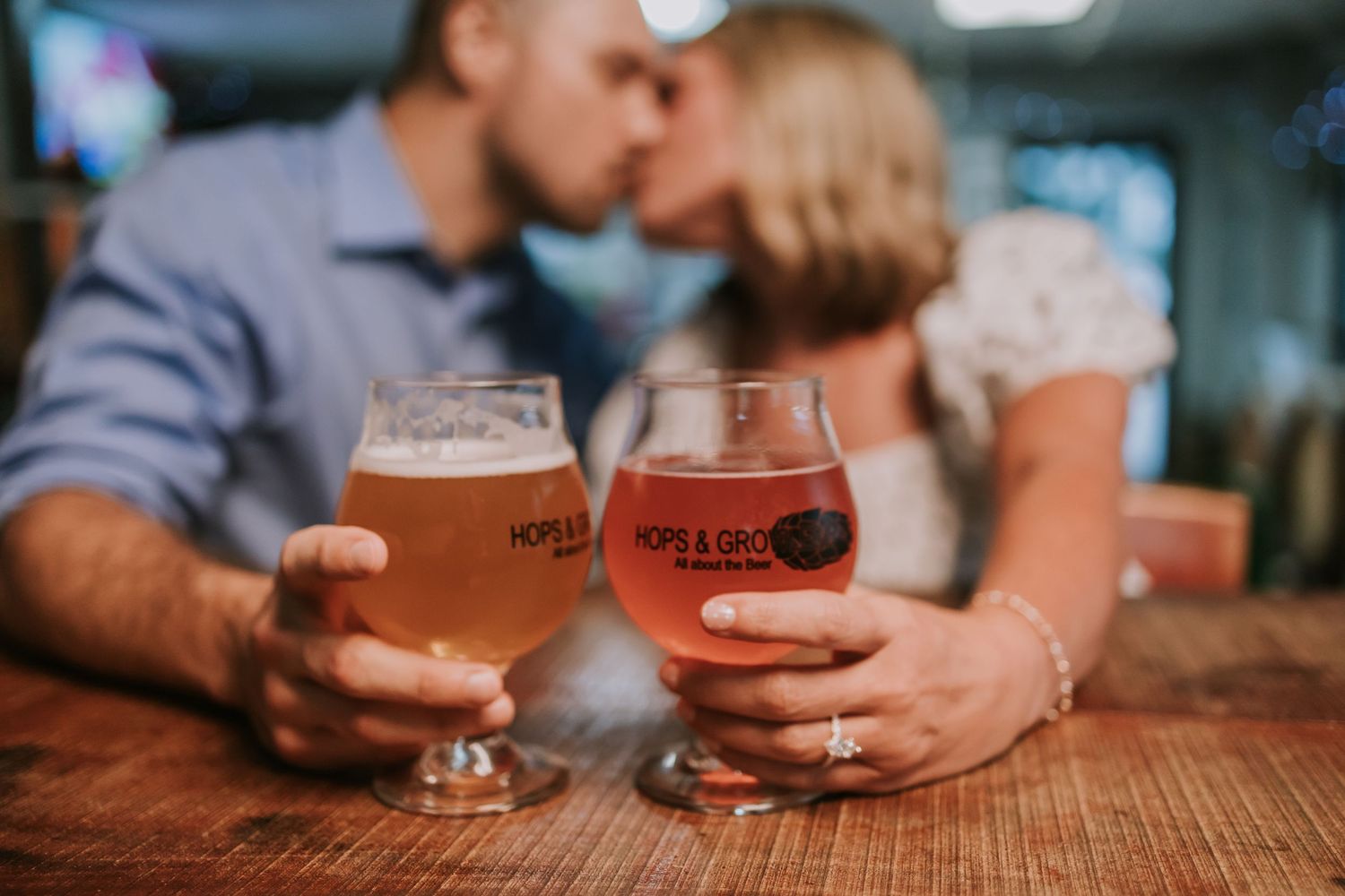 Couple kissing behind two Hops & Grains beer glasses on a wooden bar, celebrating a romantic engagement.