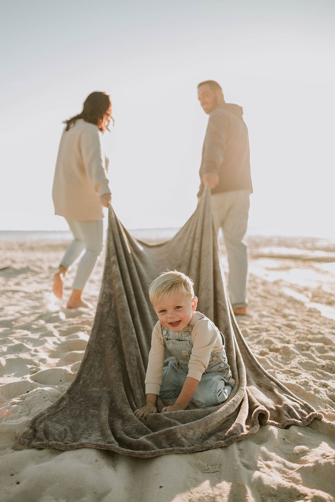 Family spending time together at the beach with a child playing in a blanket on the sand during golden hour.