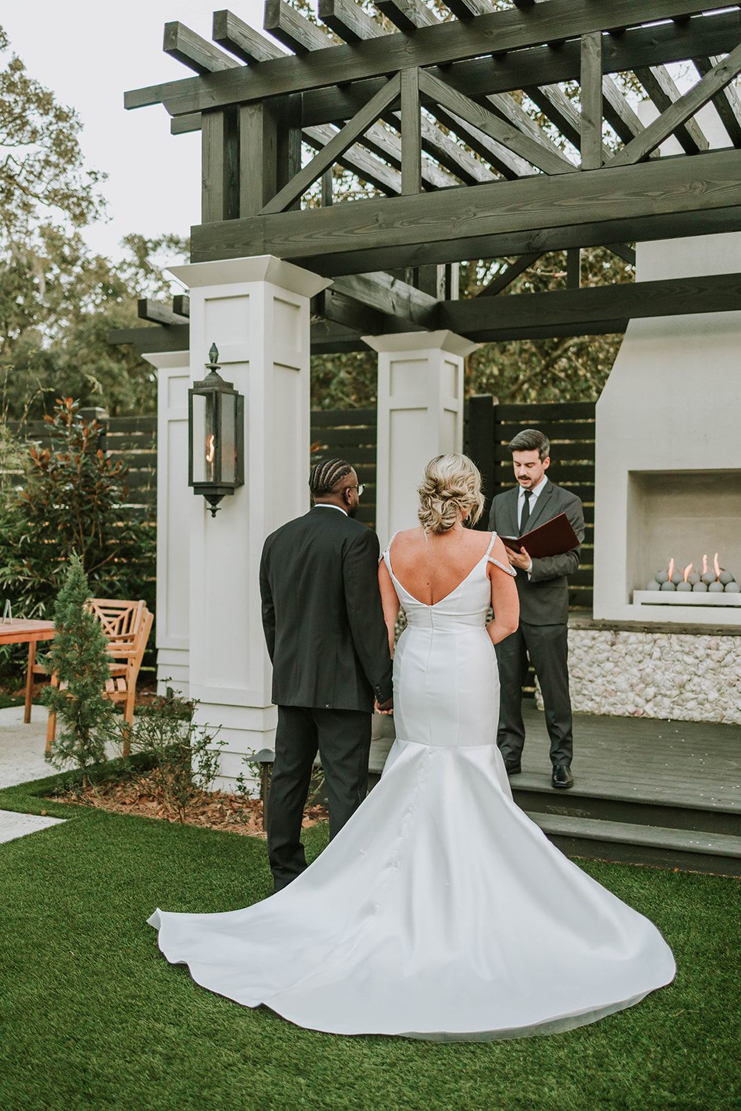 An intimate outdoor wedding ceremony under a wooden pergola on a manicured lawn with decorative lighting.